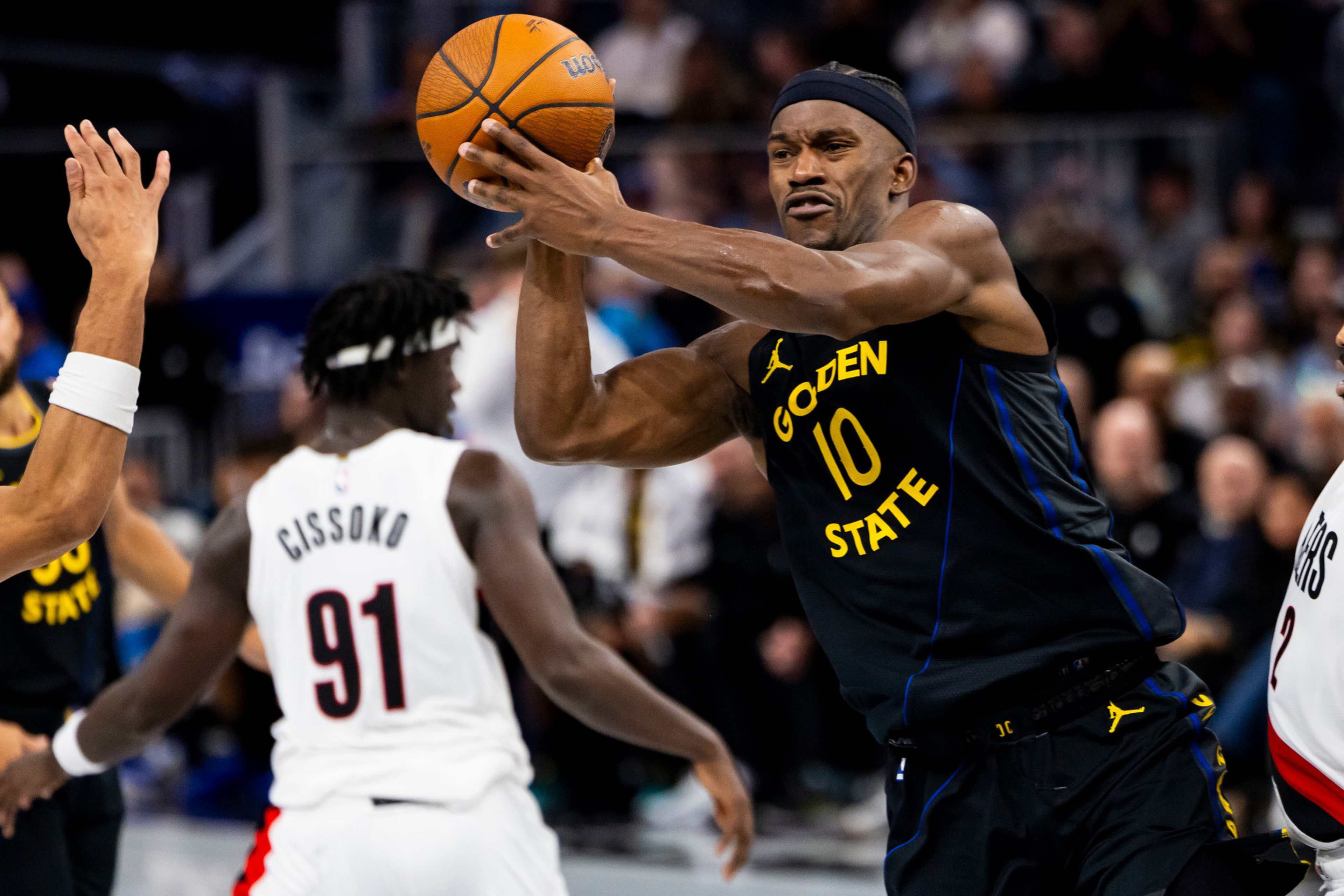 A Golden State Warriors player wearing number 10 tightly grips a basketball, preparing to pass or shoot, while a defender in a white jersey watches.