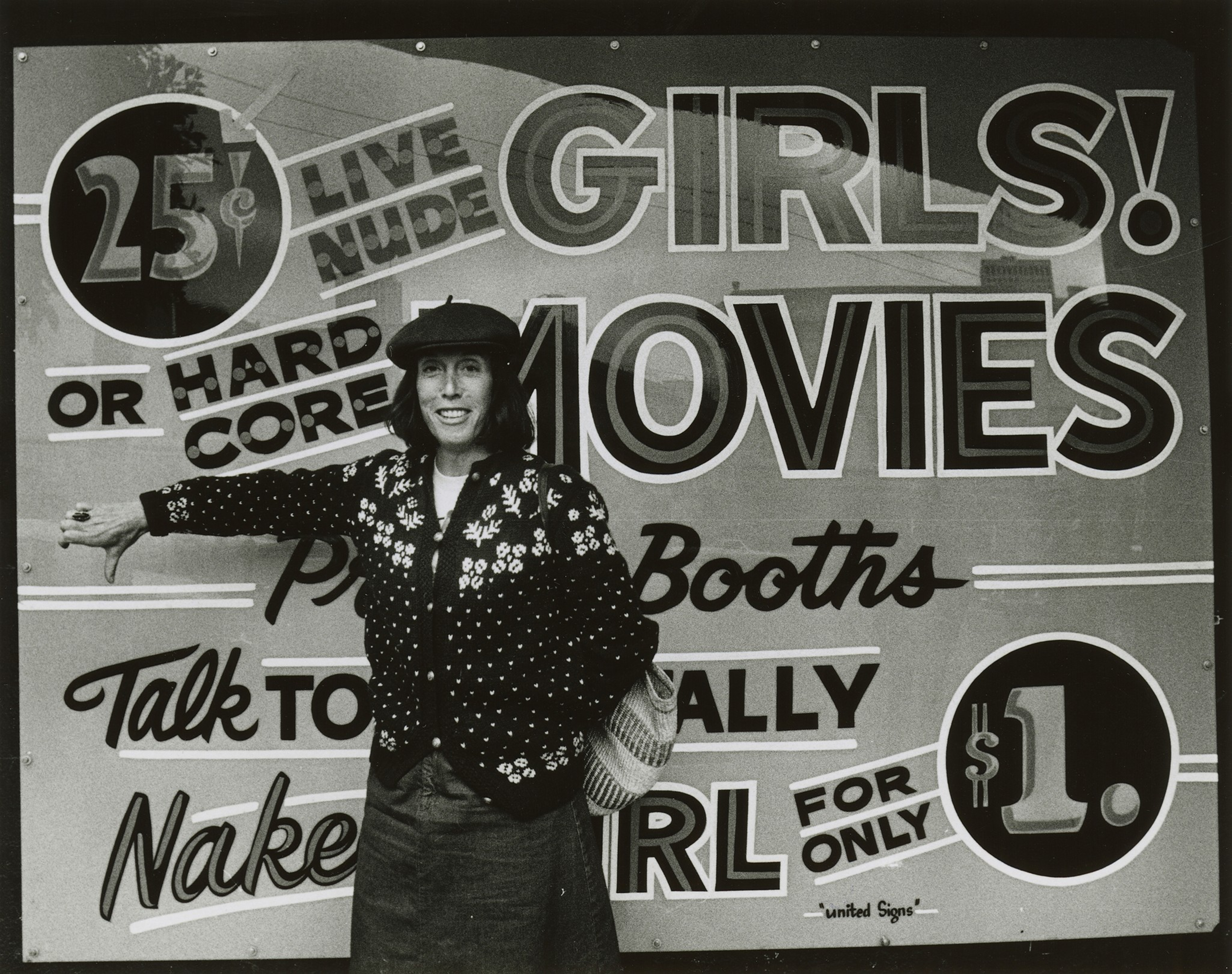 A person in a polka-dot jacket and hat stands smiling with a thumbs-down in front of a bold, colorful sign advertising "girls," "movies," and prices.