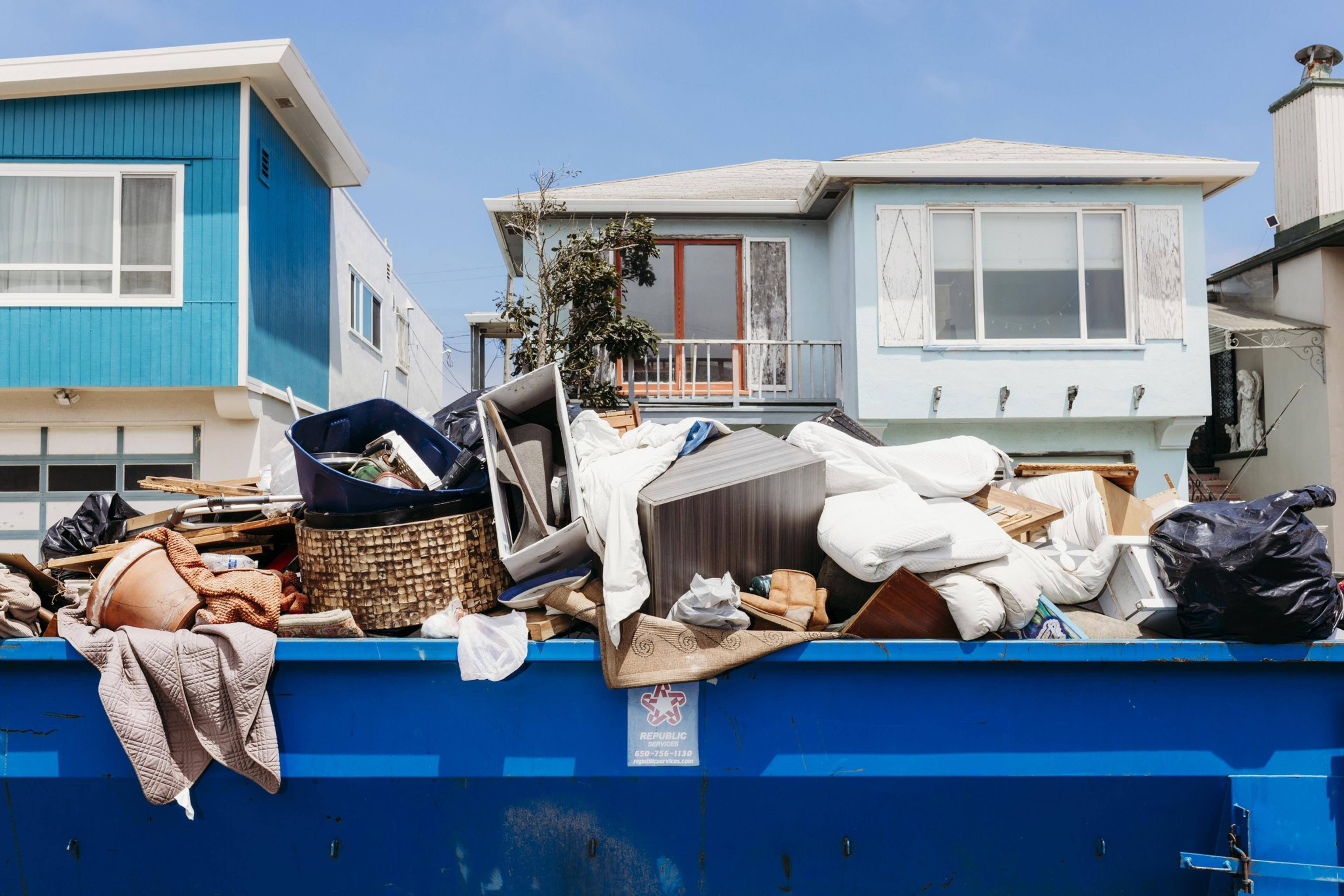 A blue dumpster overflows with household debris, including furniture, mattresses, and bags, in front of two adjacent houses, one blue and one light blue.