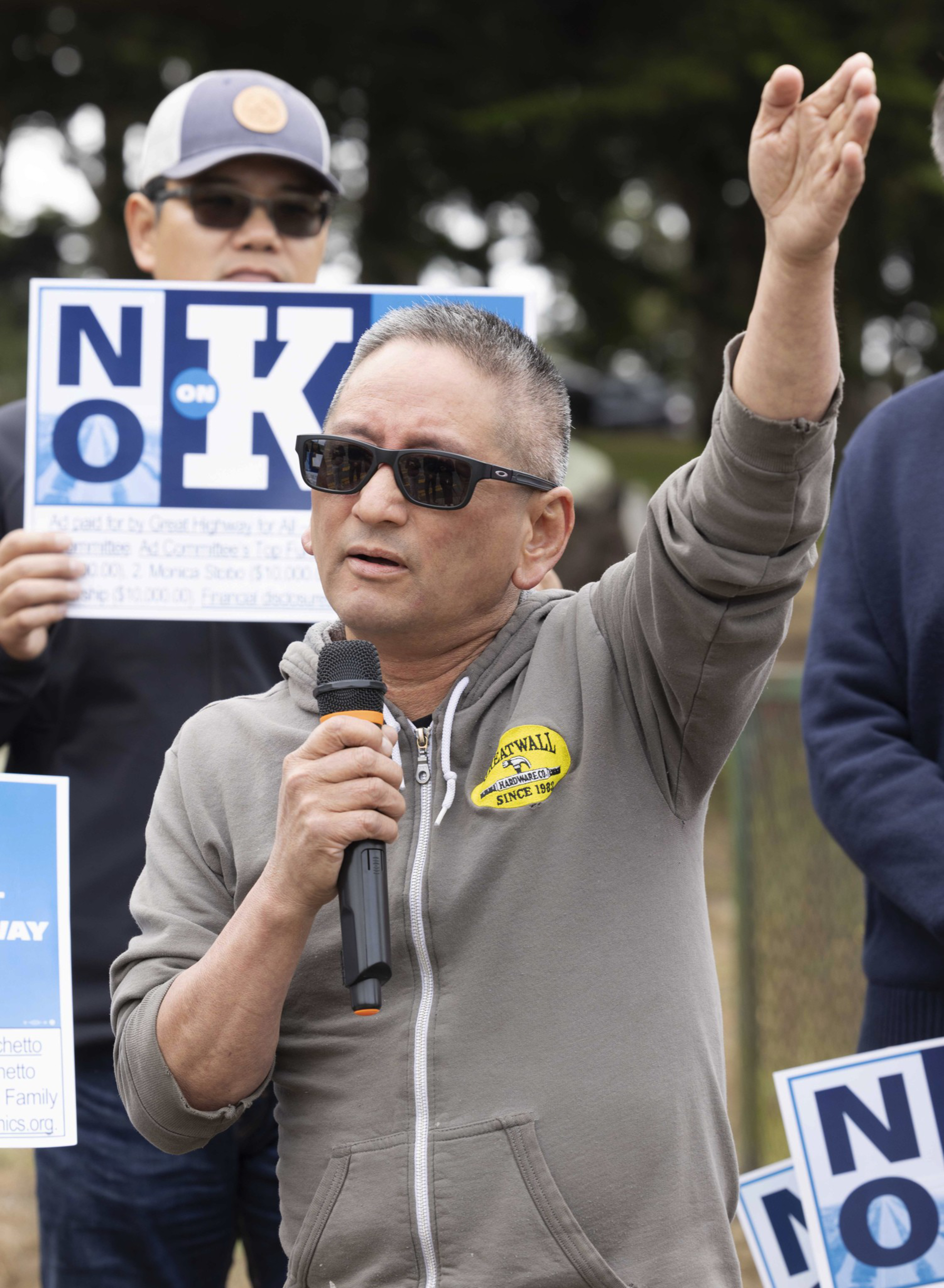 A man in sunglasses and a gray hoodie speaks into a microphone, gesturing with one hand. Behind him, another person holds a &quot;No on K&quot; sign.