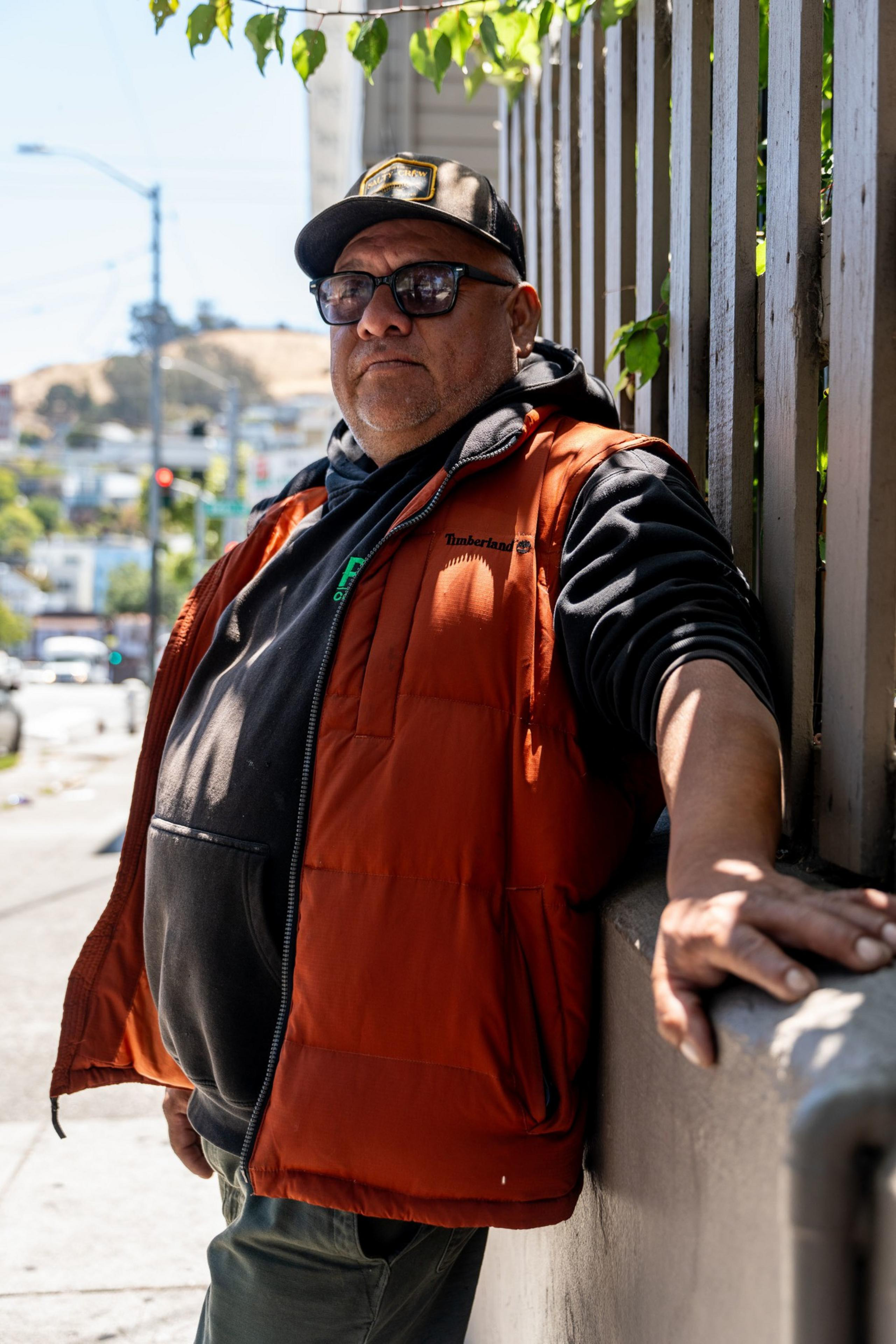 A man leans on a railing outdoors, wearing sunglasses, a black cap, an orange vest, and a black hoodie. The sun casts shadows, and trees are in the background.