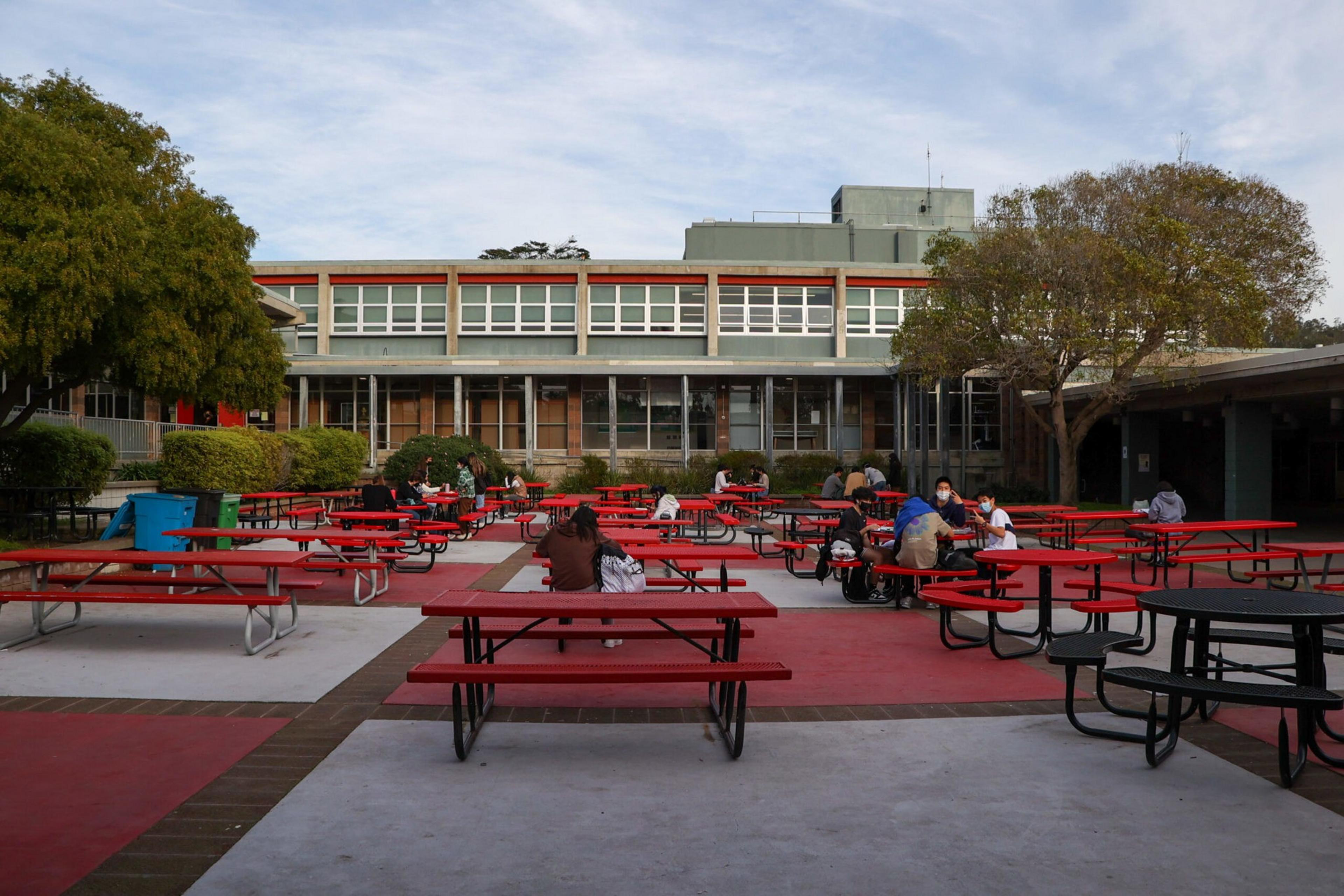 Outdoor courtyard with red and black picnic tables, several people sitting at tables, surrounded by trees and a two-story building.