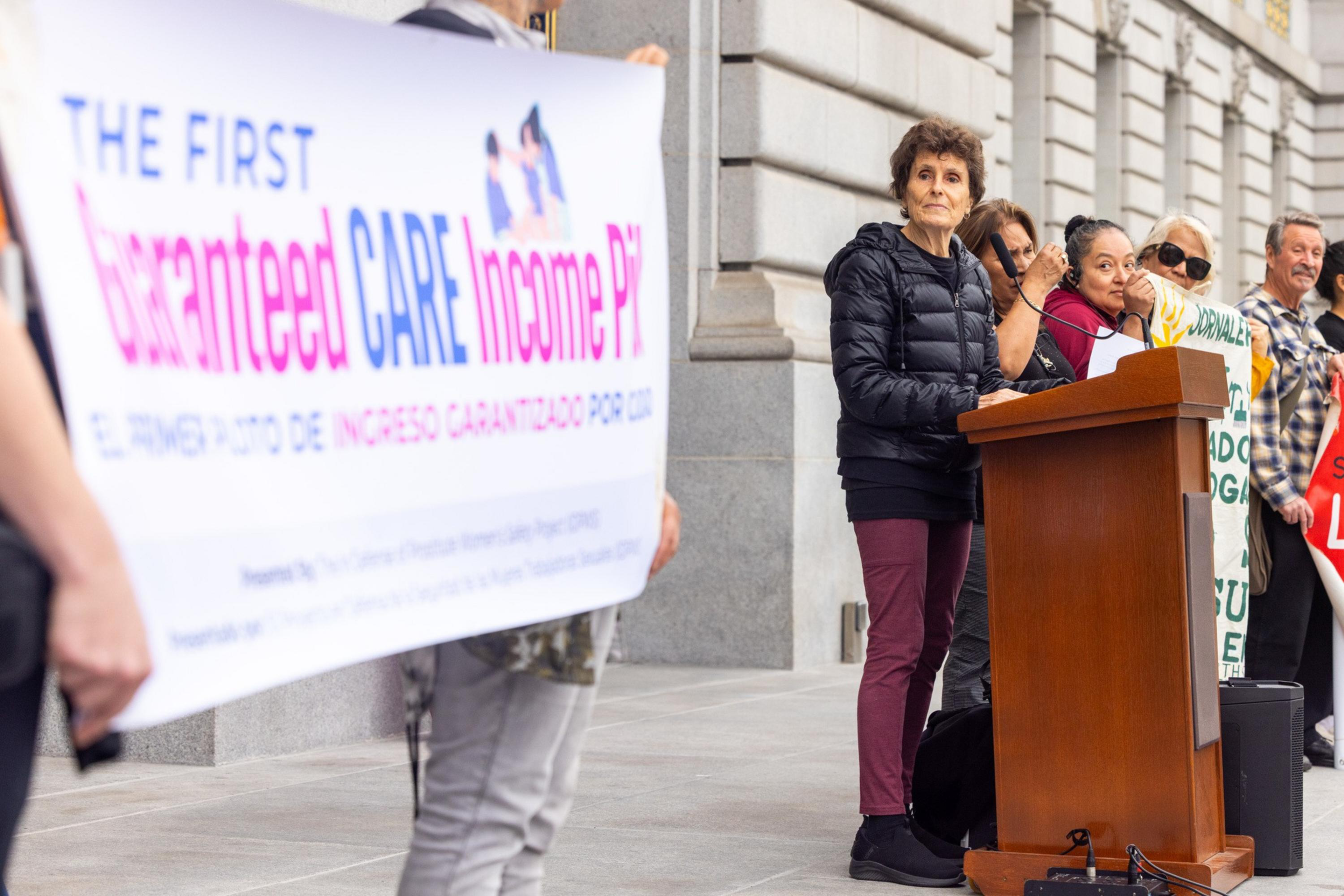 A woman speaks at a podium while others hold signs advocating for guaranteed CARE income payments outside a stone building.