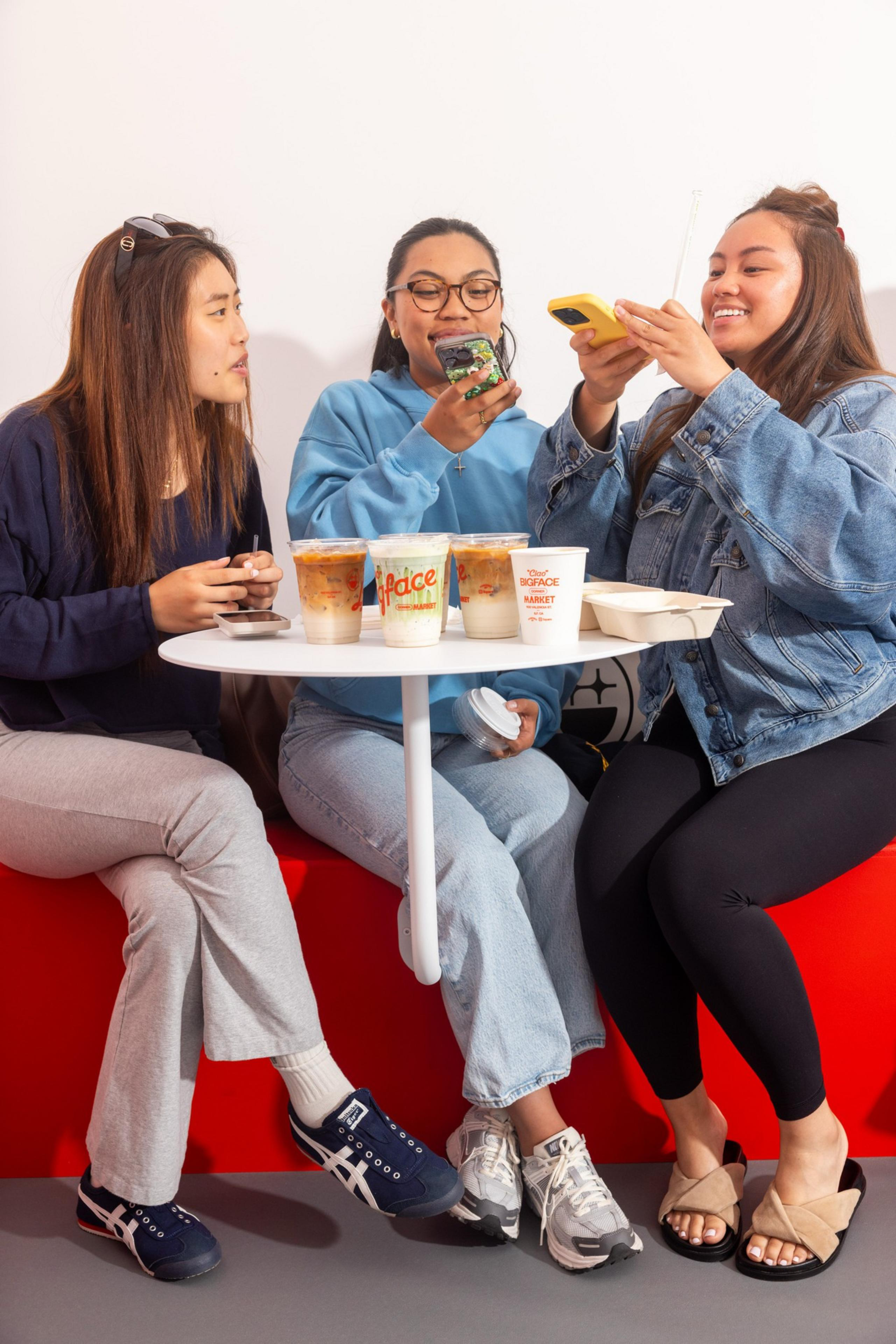 Three people are sitting around a white table, sharing drinks and using their phones. They appear relaxed and happy, seated on a red bench.