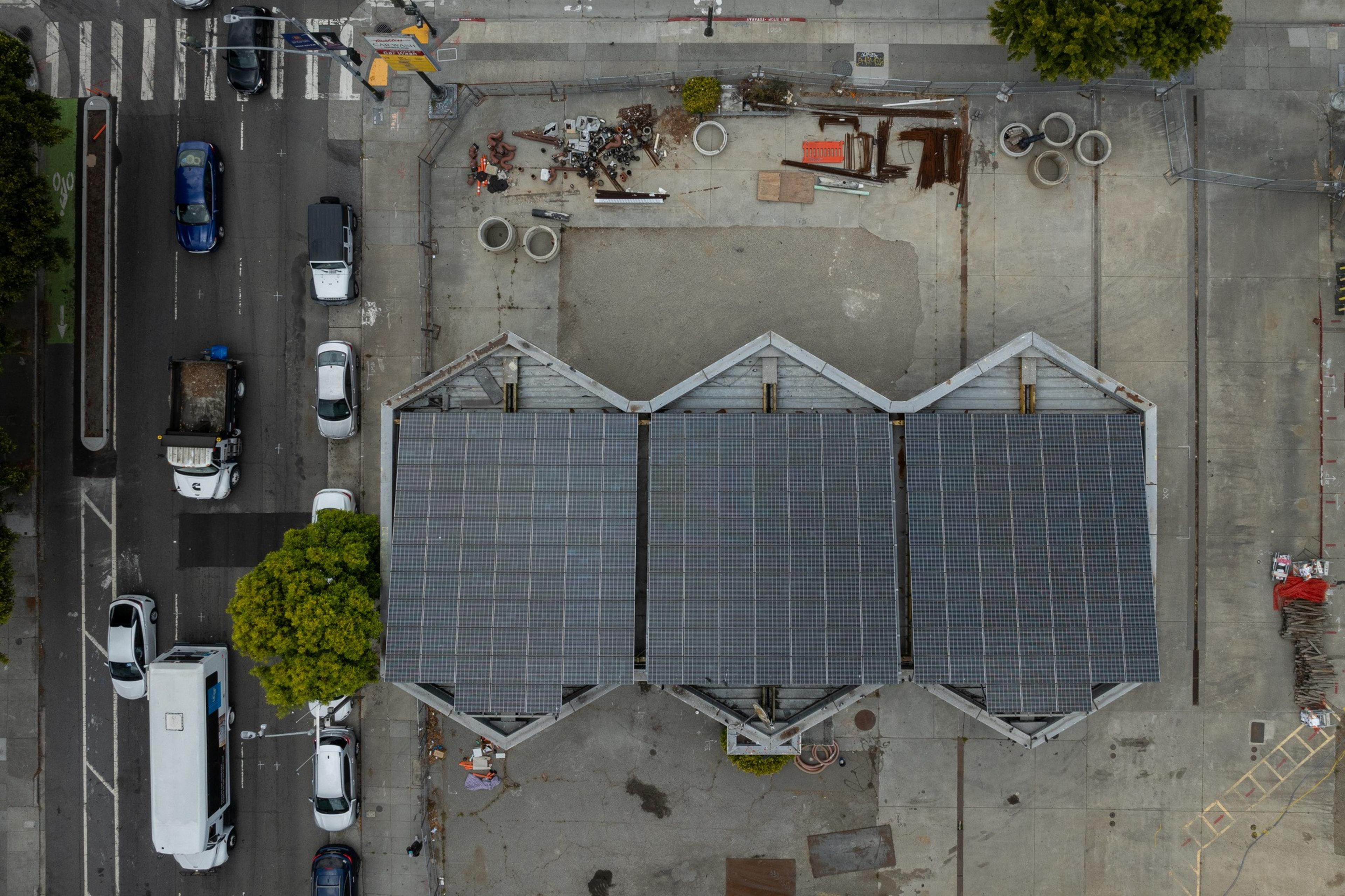 The image shows an overhead view of a construction site, with a large building roof covered in solar panels, nearby parked cars, and scattered construction materials.