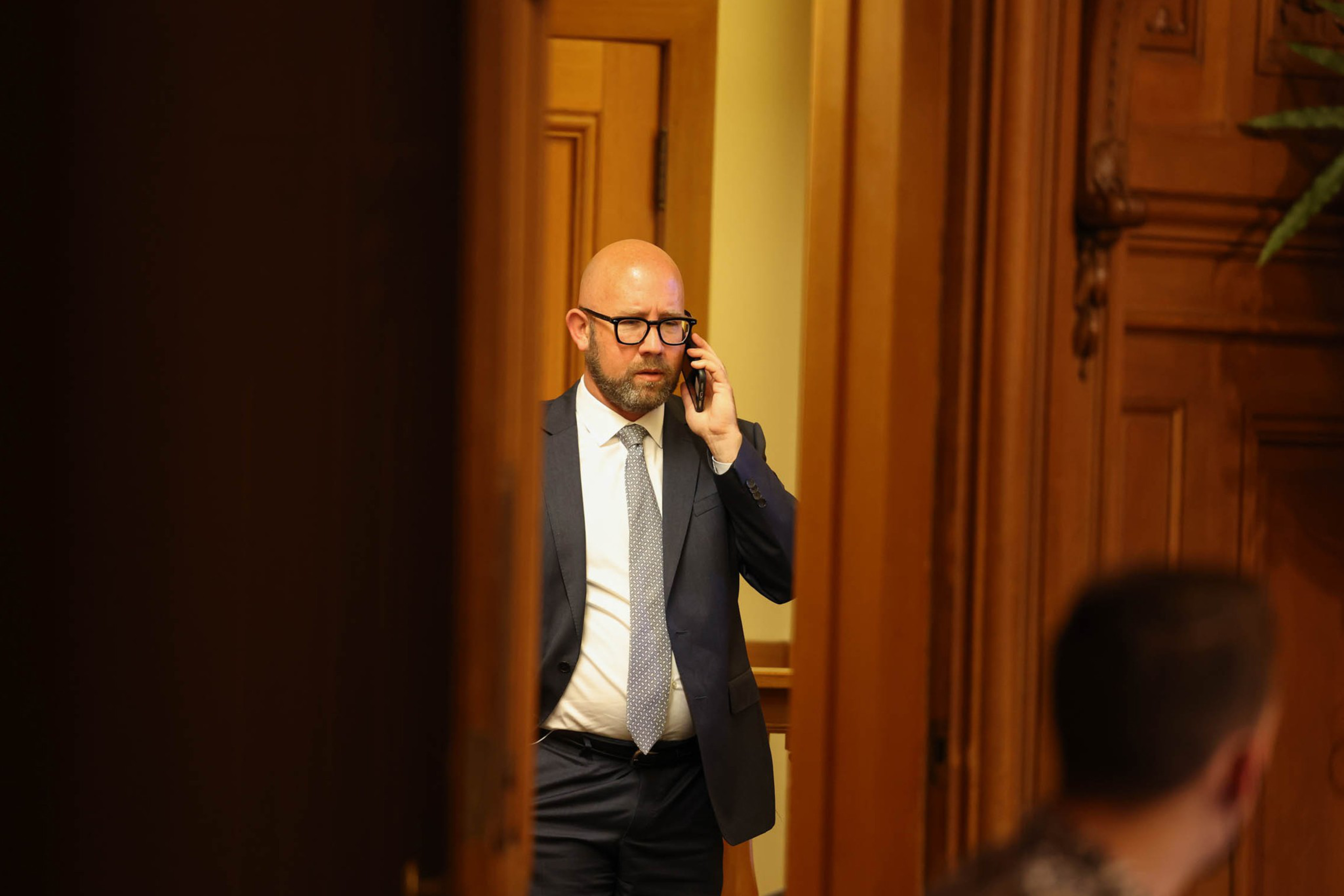 A bald man in glasses and a suit talks on a phone in a wood-paneled room, seen through a partially open door.
