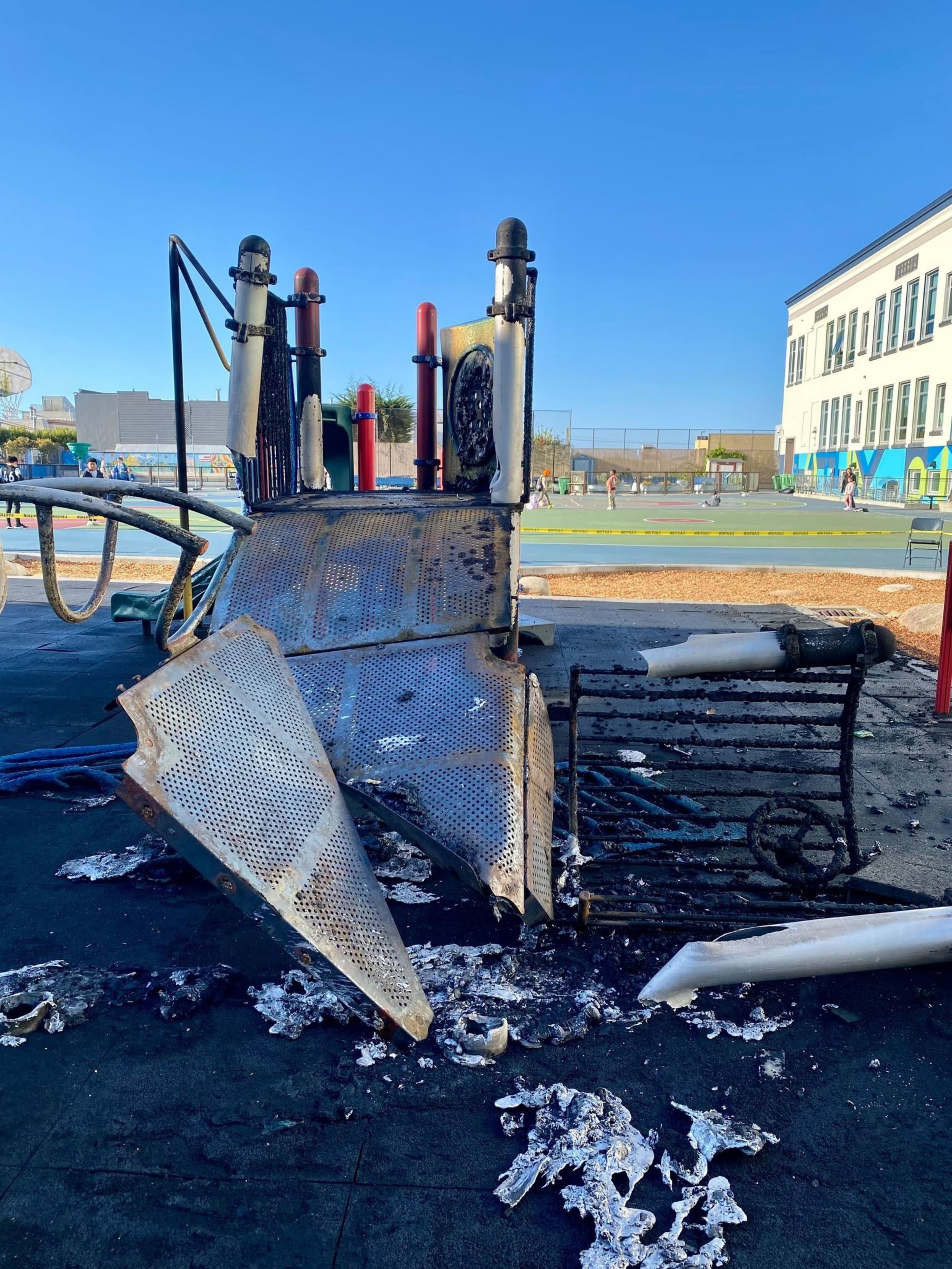 The image shows a burnt playground slide with melted plastic and metal debris. A school building and basketball court are visible in the background.