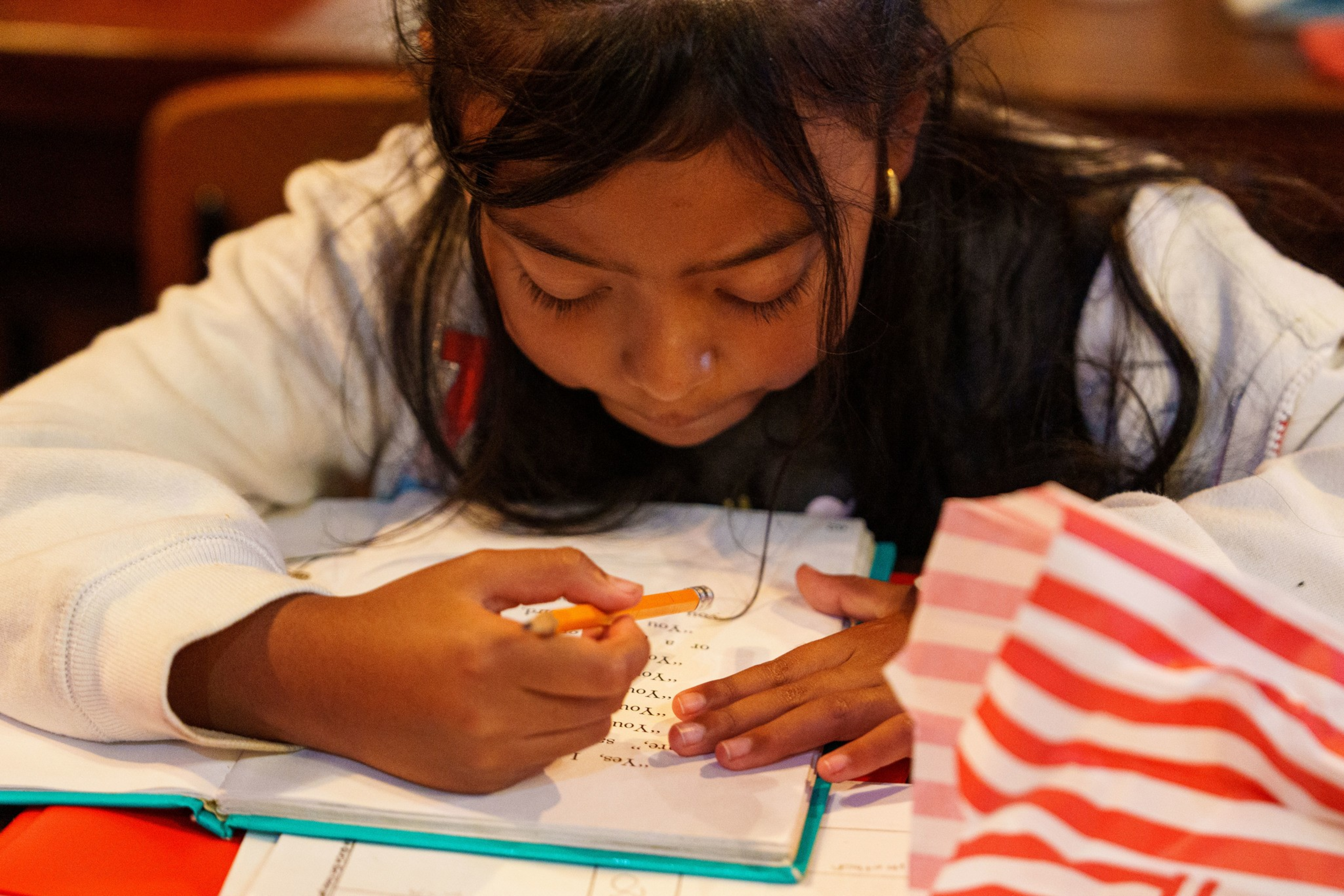 A young girl with long dark hair is focused on writing in a notebook with a pencil while resting her arms on a table.