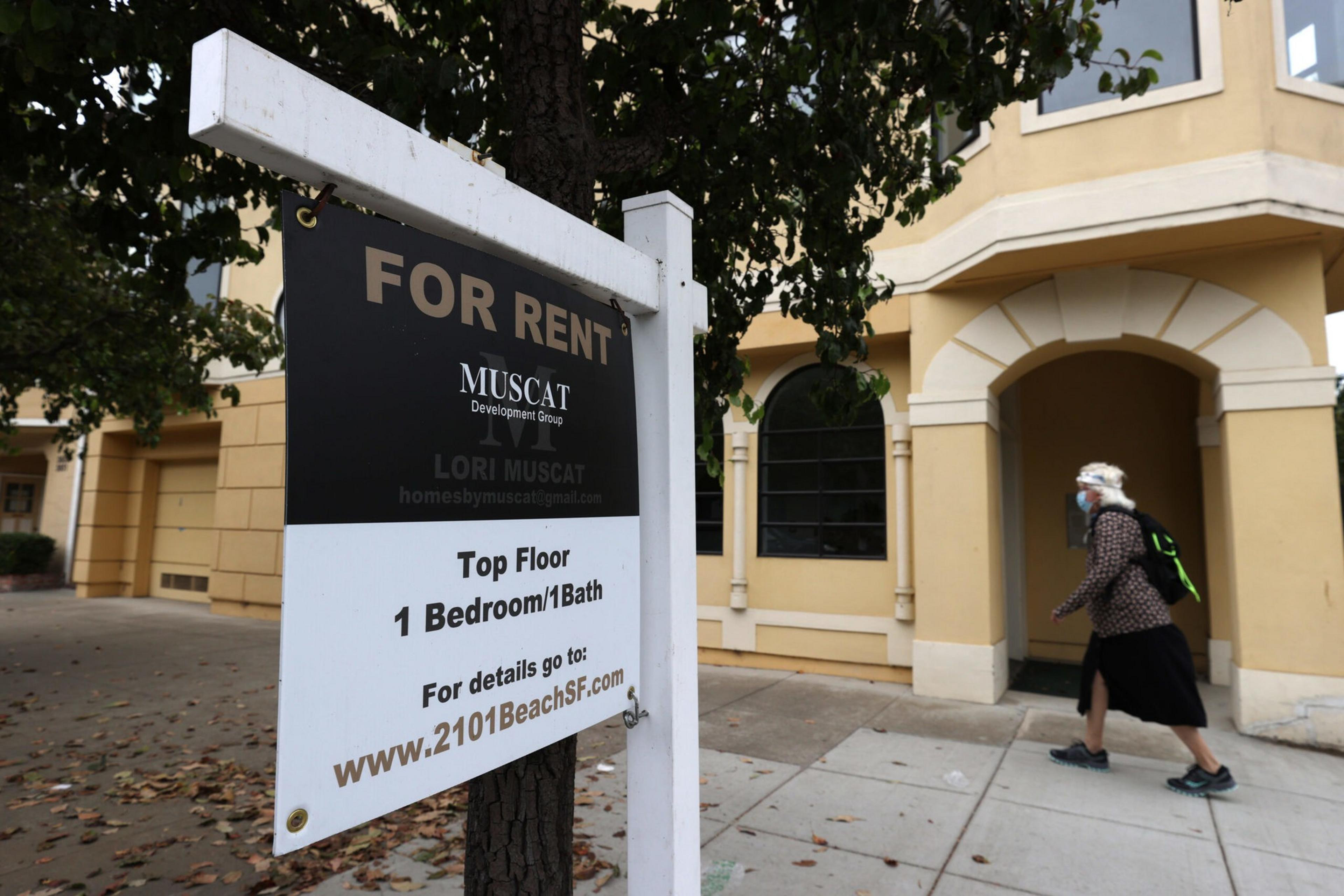 A "For Rent" sign offers a top floor 1-bedroom, 1-bath apartment, with a person wearing a mask and backpack walking past a beige building.