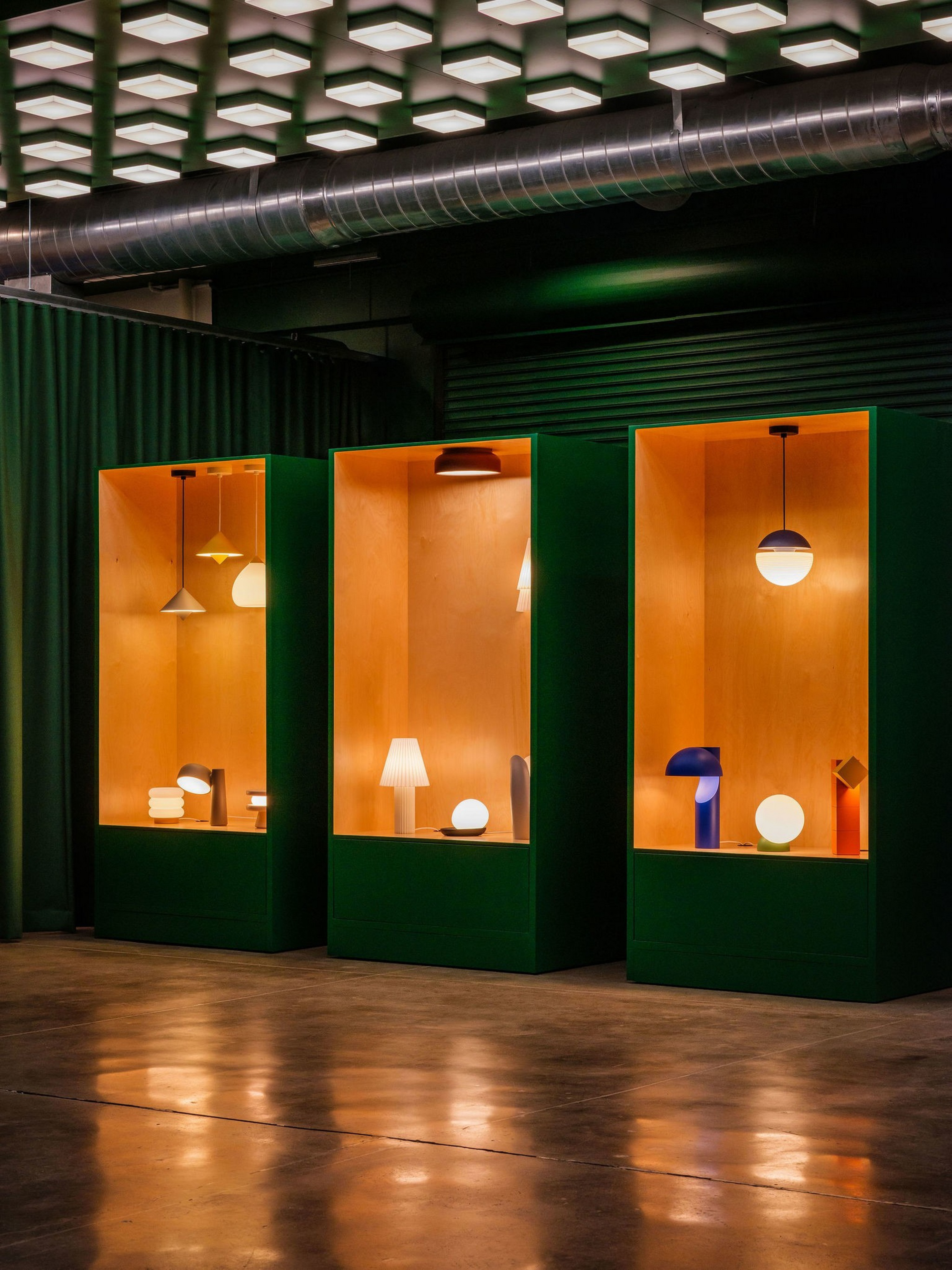Three green display cases with wooden interiors each showcase a variety of illuminated modern lamps and light fixtures against a dark backdrop.