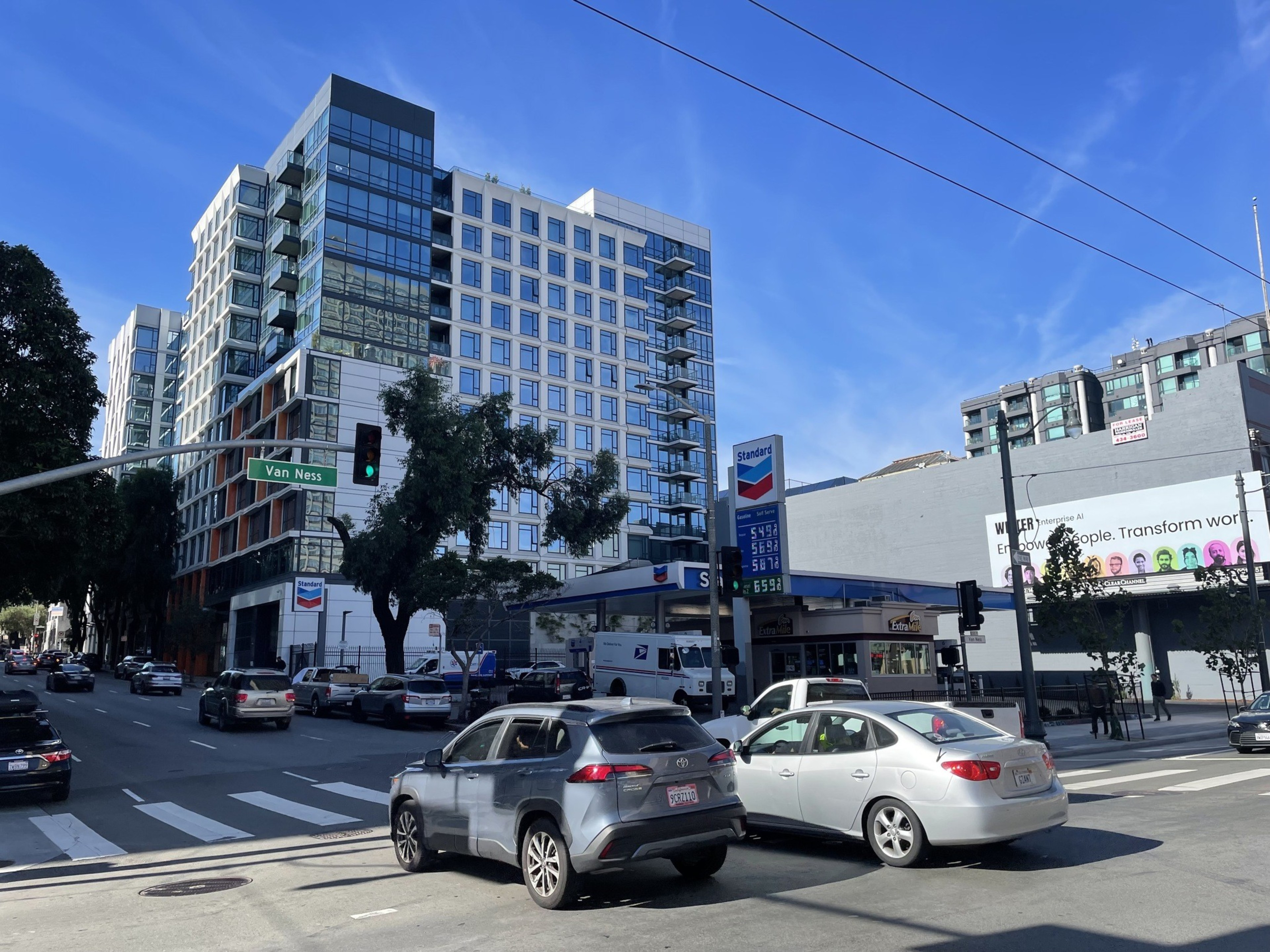 Cars drive through an intersection past a gas station with a large condo building in the background.
