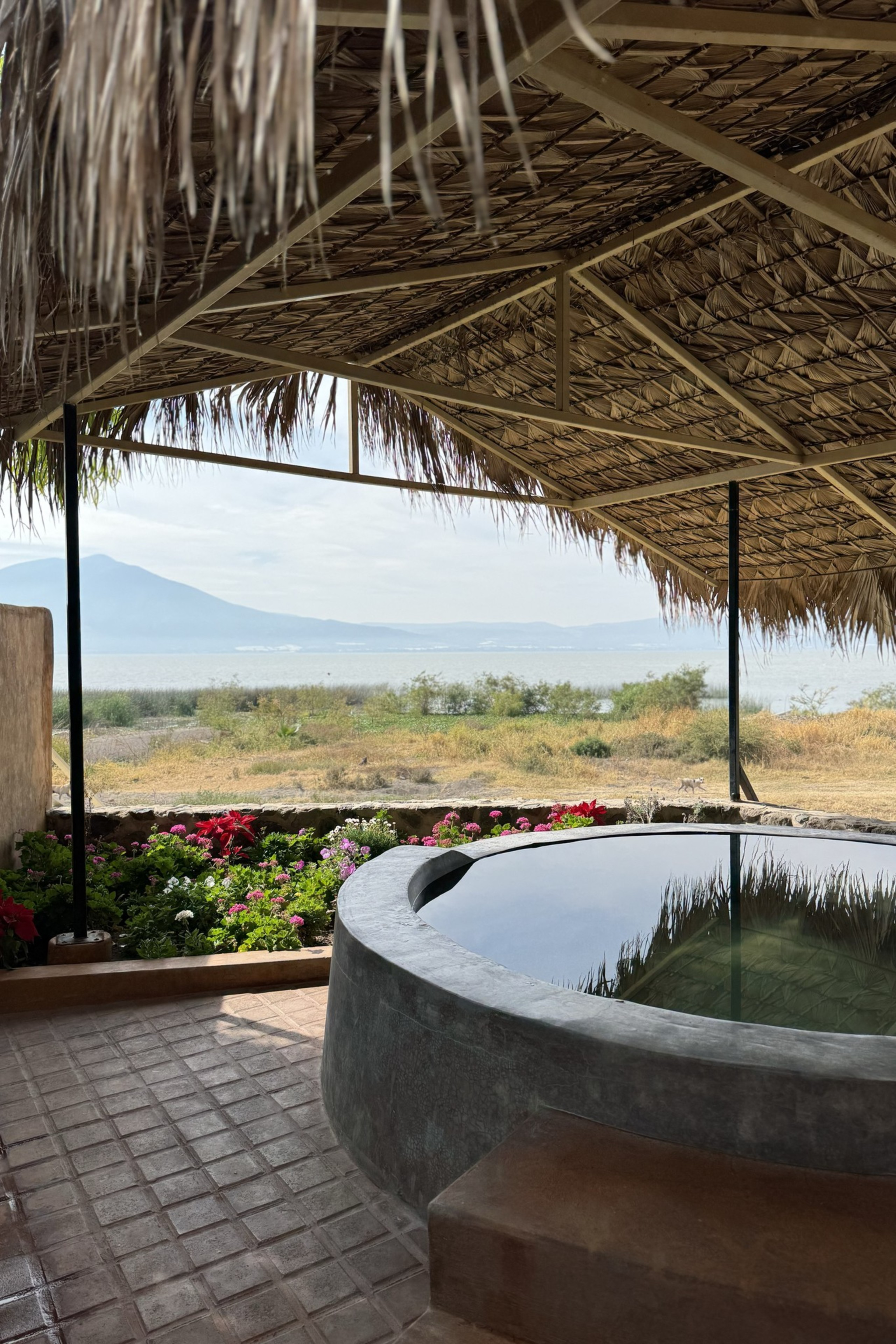 The image shows a round hot tub under a thatched roof, overlooking a lake with mountains in the distance. There are colorful flowers and grass nearby.
