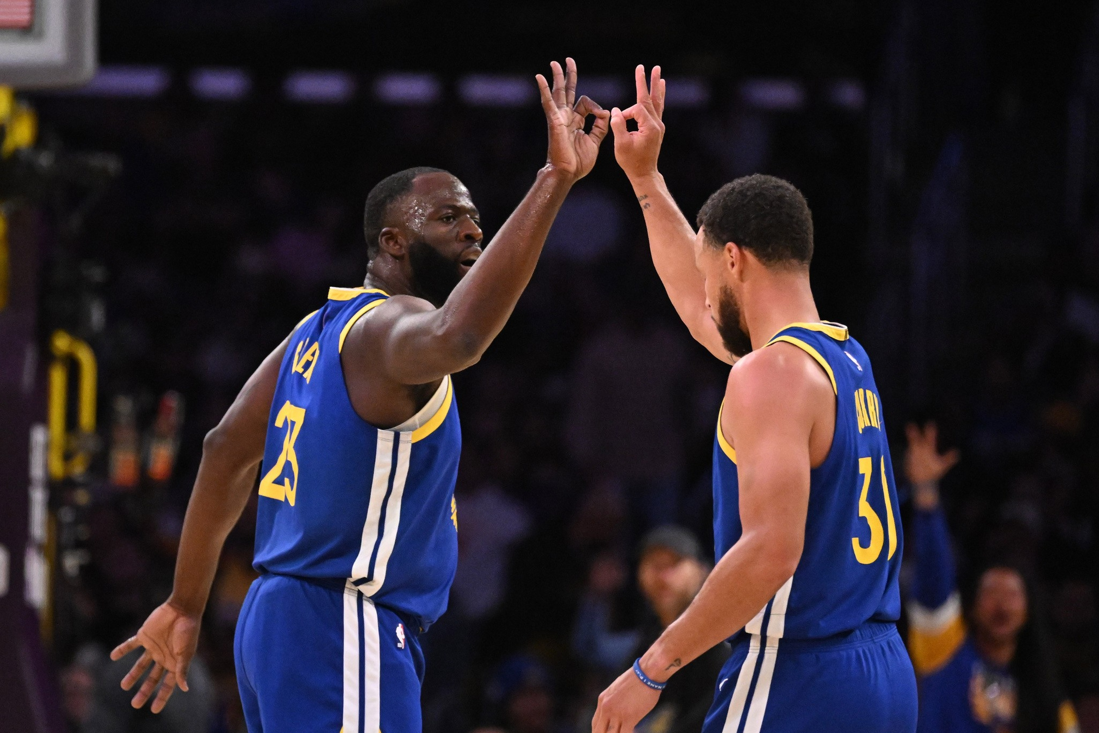 Two basketball players in blue and yellow uniforms are giving each other a high-five during a game, with one player facing forward and the other backward.