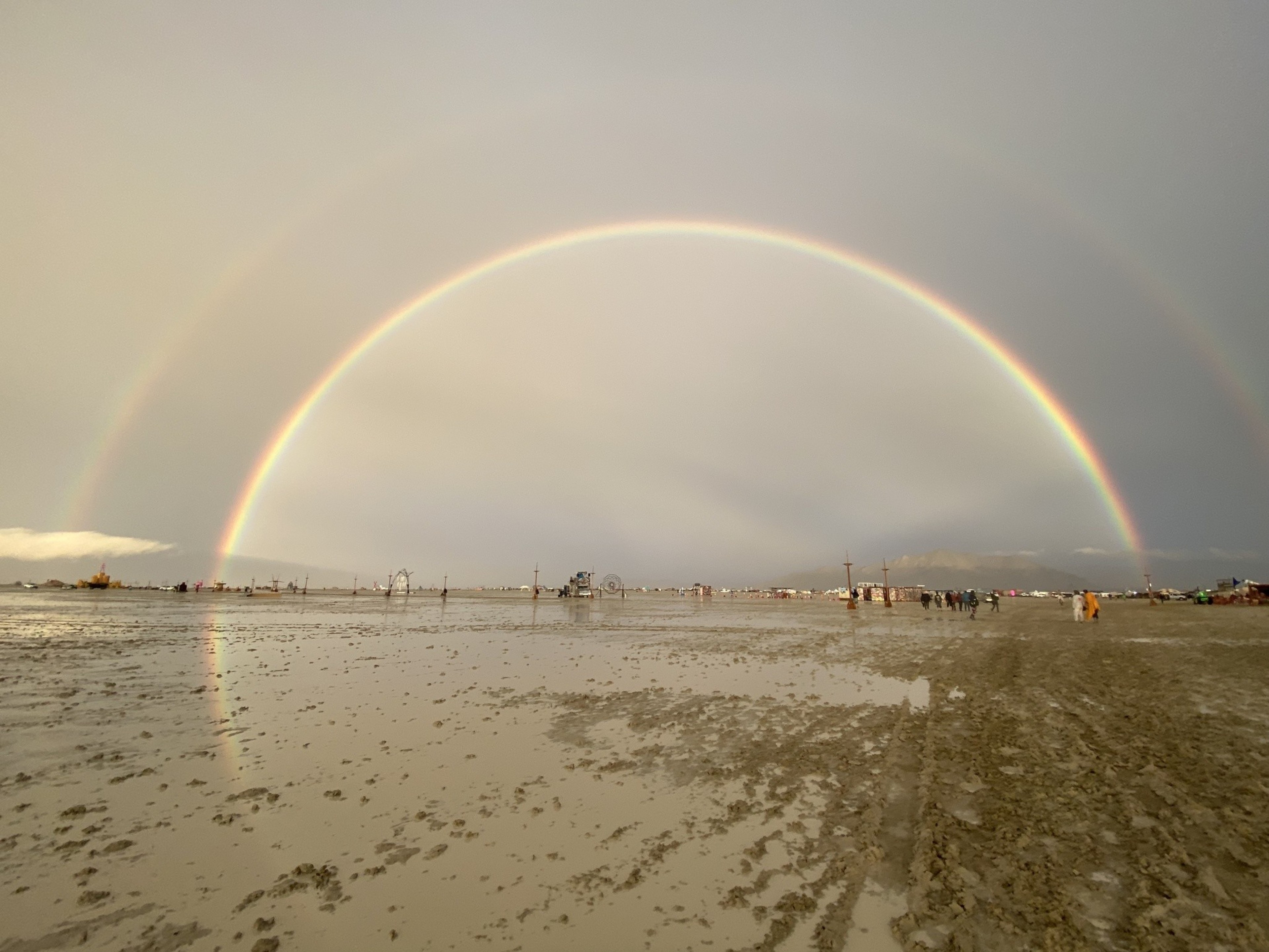 A double rainbow arches over a wet, muddy landscape with scattered people and structures.