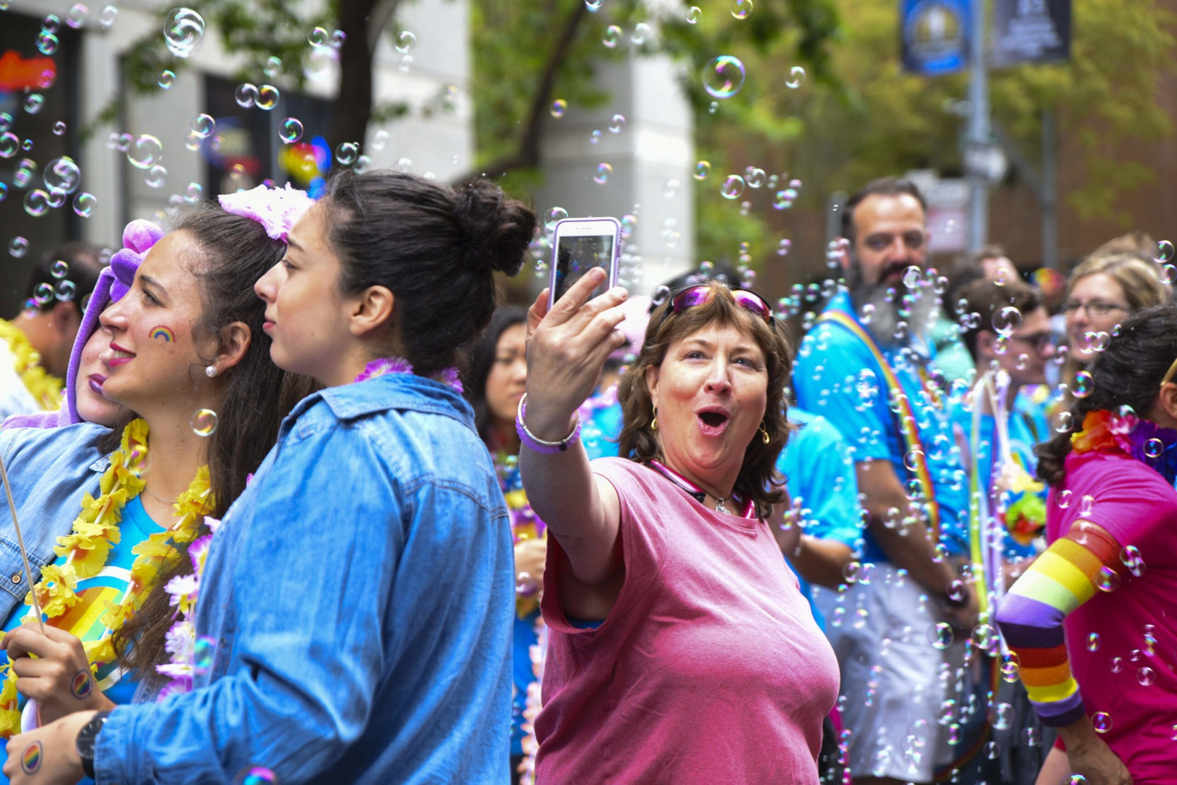 People at a colorful outdoor event surrounded by bubbles; a woman in pink takes a selfie while others smile and wear rainbow-themed accessories.