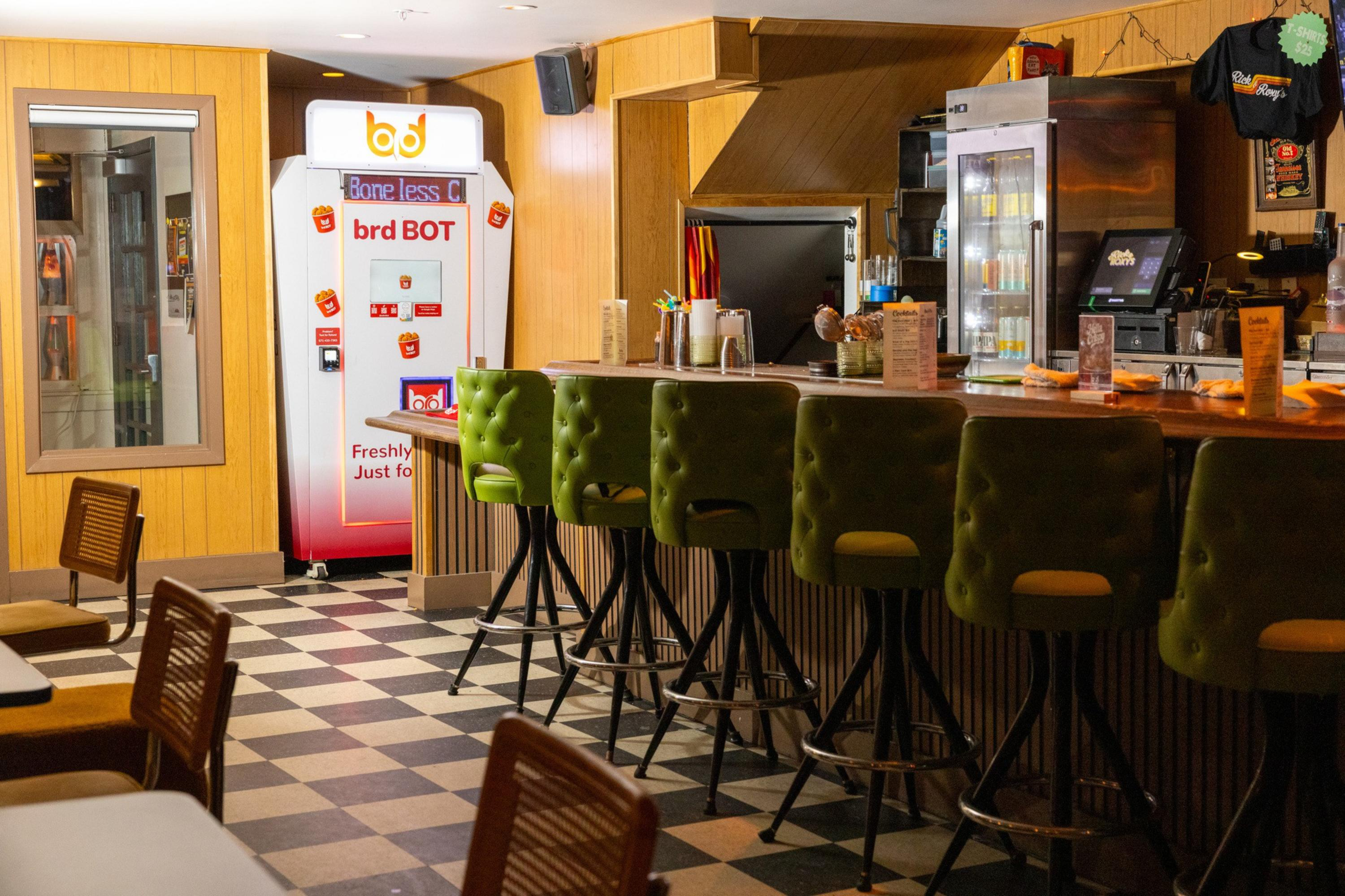 A retro-style diner with a checkered floor, green cushioned bar stools, wooden panel walls, a refrigerated drink display, and a vending machine labeled "brd BOT."
