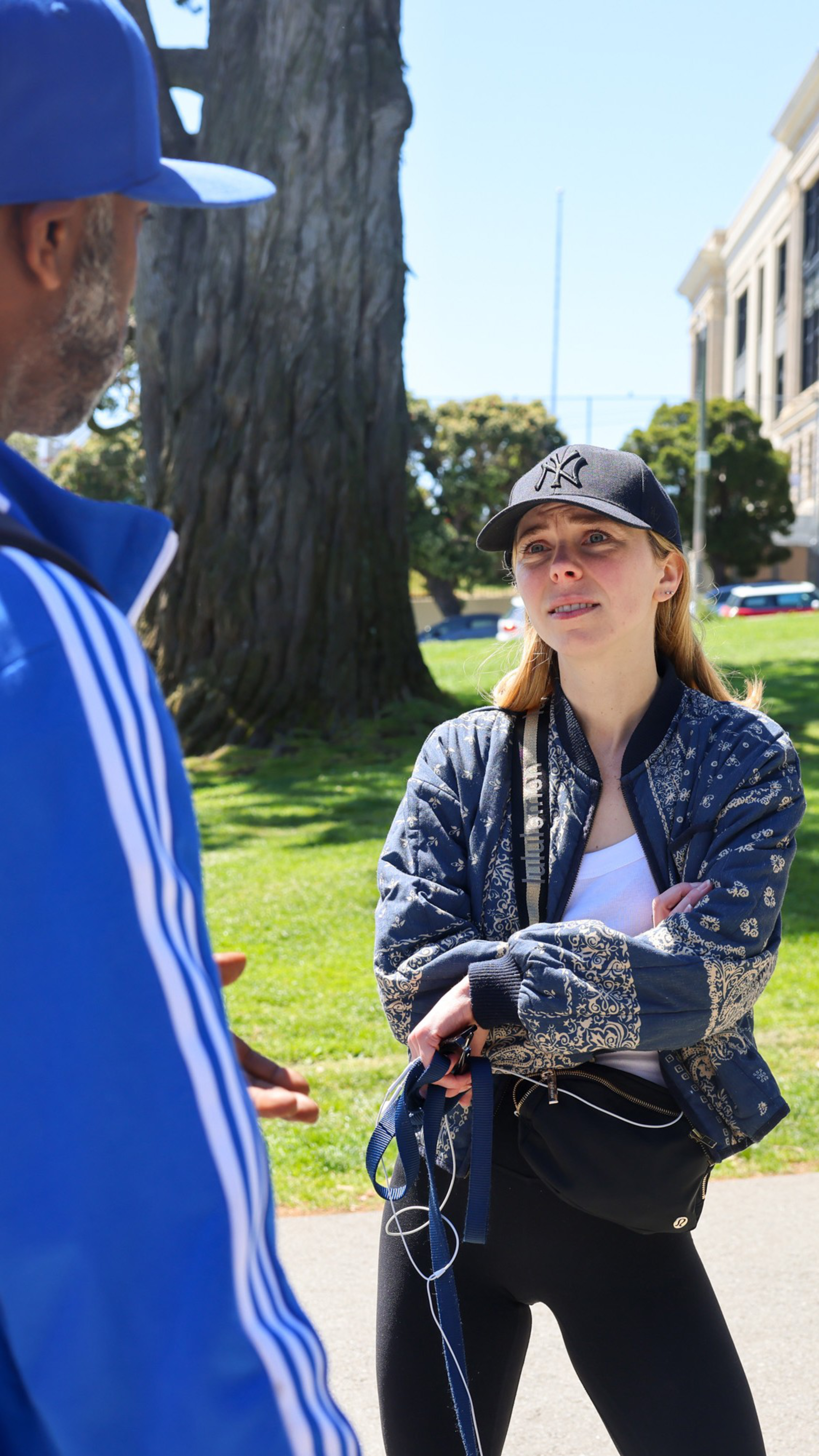 A person in a Yankees cap listens intently to a man in a blue cap, with a tree and clear sky in the background.