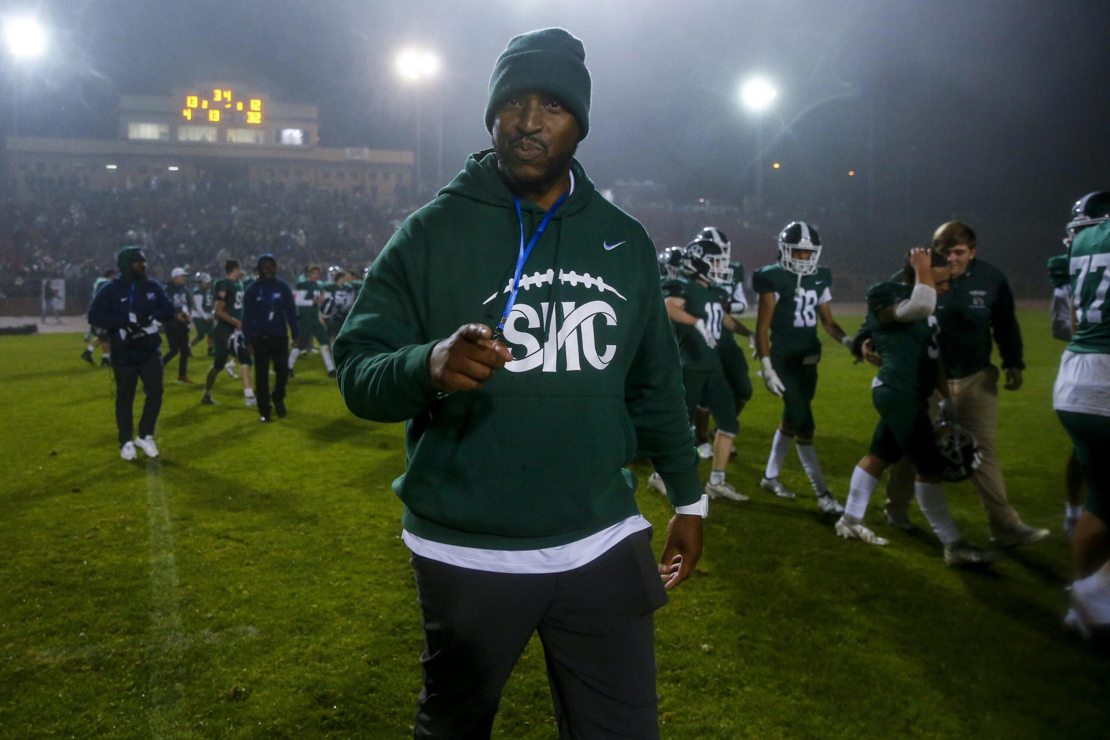 A football coach in a green hoodie and beanie points toward the camera on a foggy, brightly lit field with players and spectators in the background.