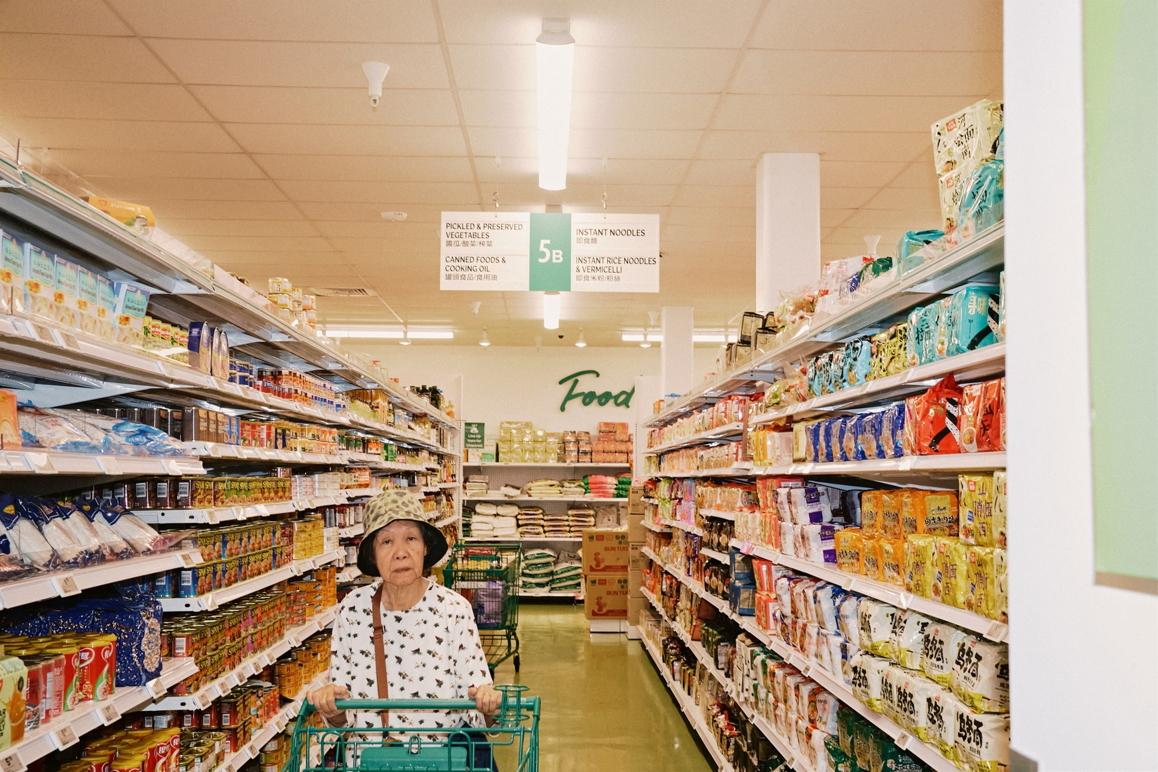 An elderly woman wearing a patterned hat and shirt pushes a shopping cart down a grocery aisle stocked with various packaged food products.