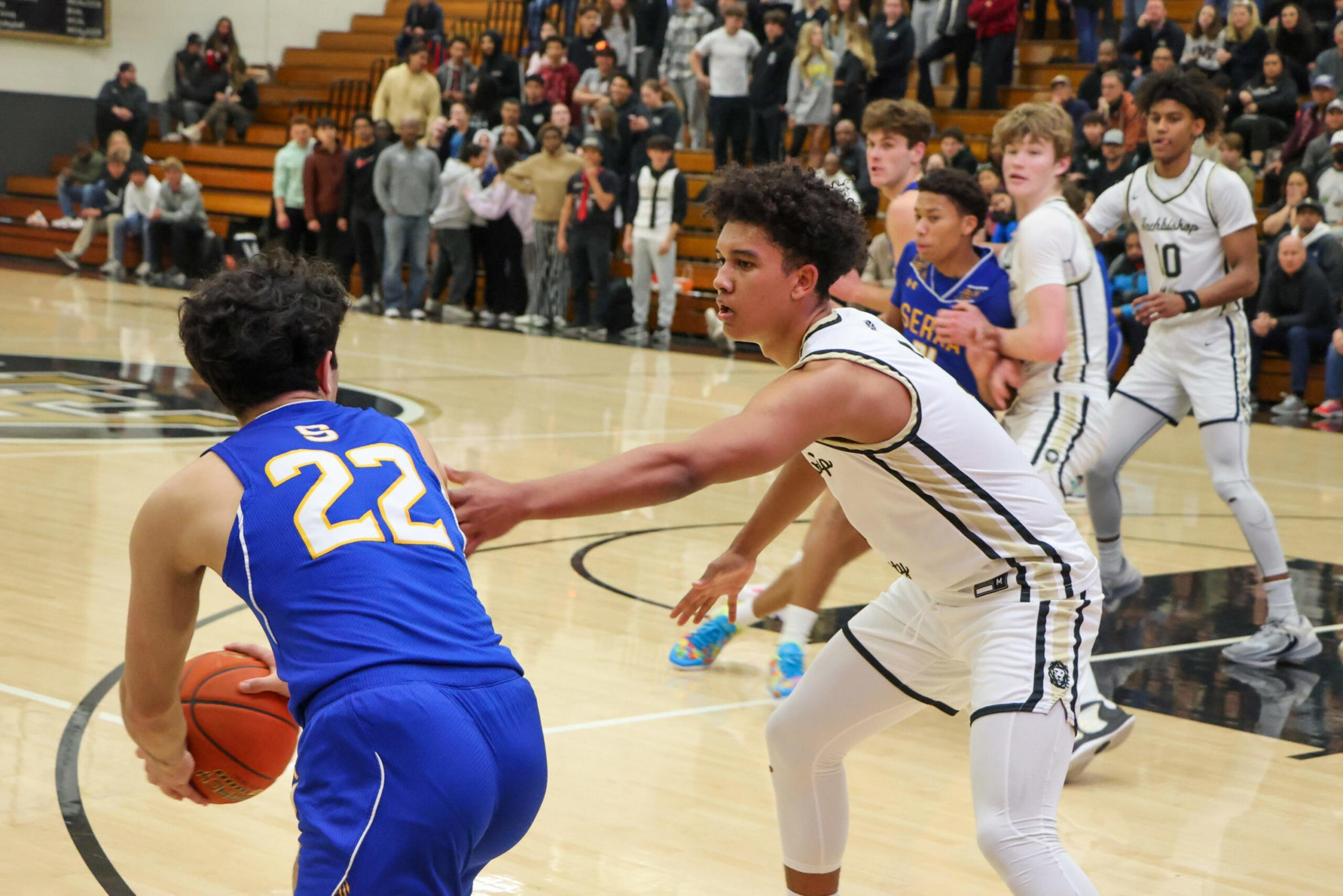 A basketball player in blue dribbles the ball while a defender in white reaches out, with other players and spectators watching in the gym.