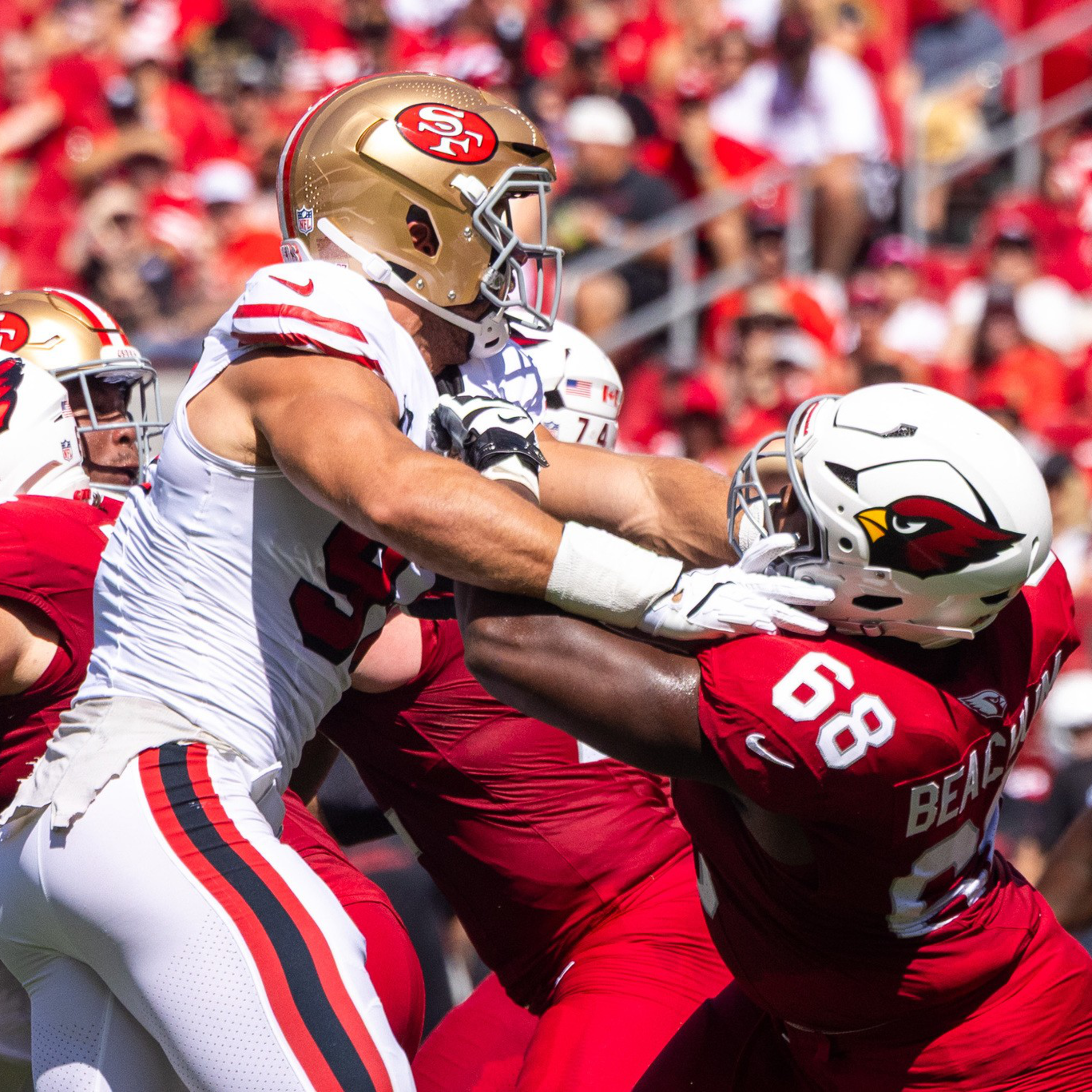 A San Francisco 49ers player in white blocks a struggling Arizona Cardinals player in red during a football game with a crowd in the background.