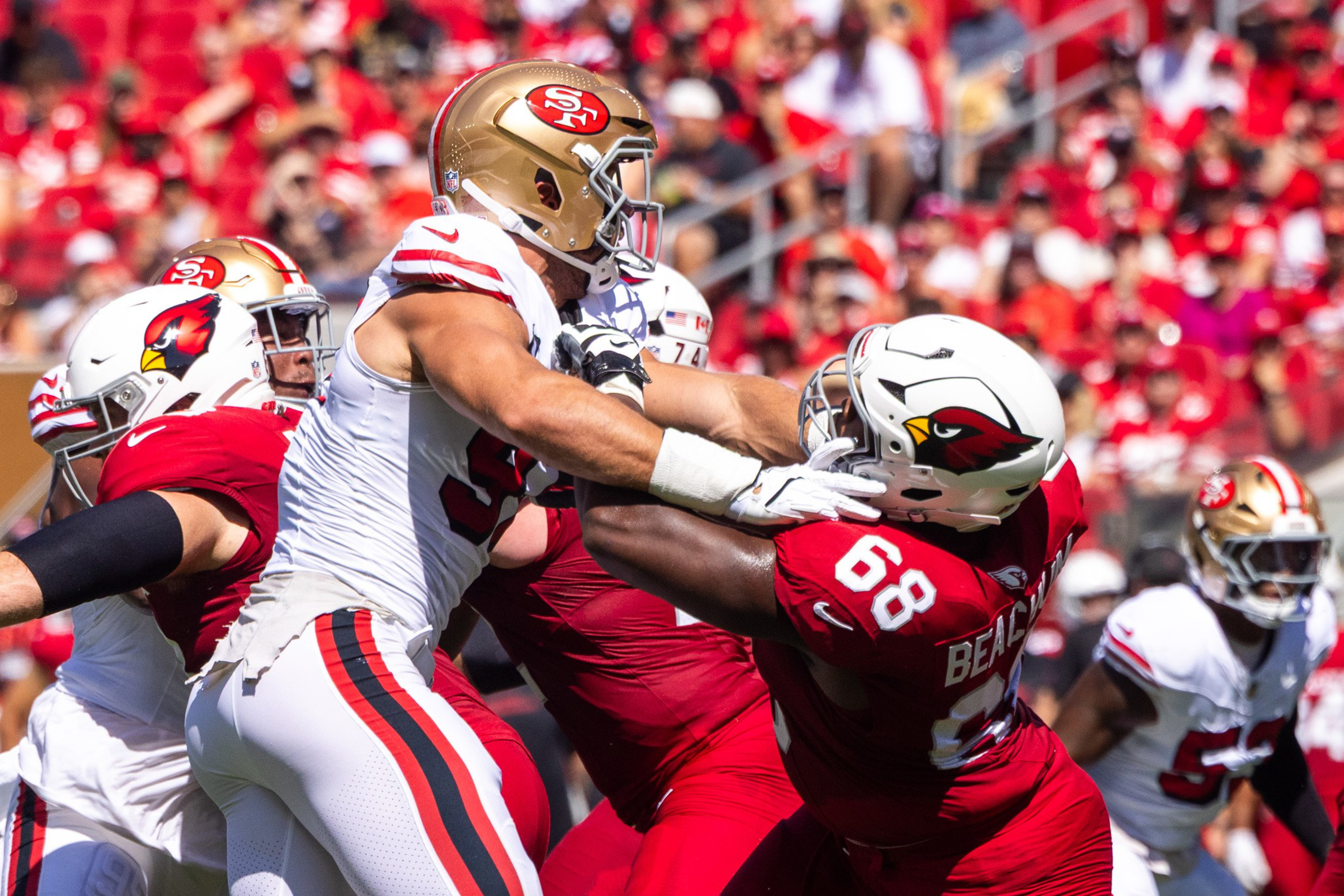 A San Francisco 49ers player in white blocks a struggling Arizona Cardinals player in red during a football game with a crowd in the background.