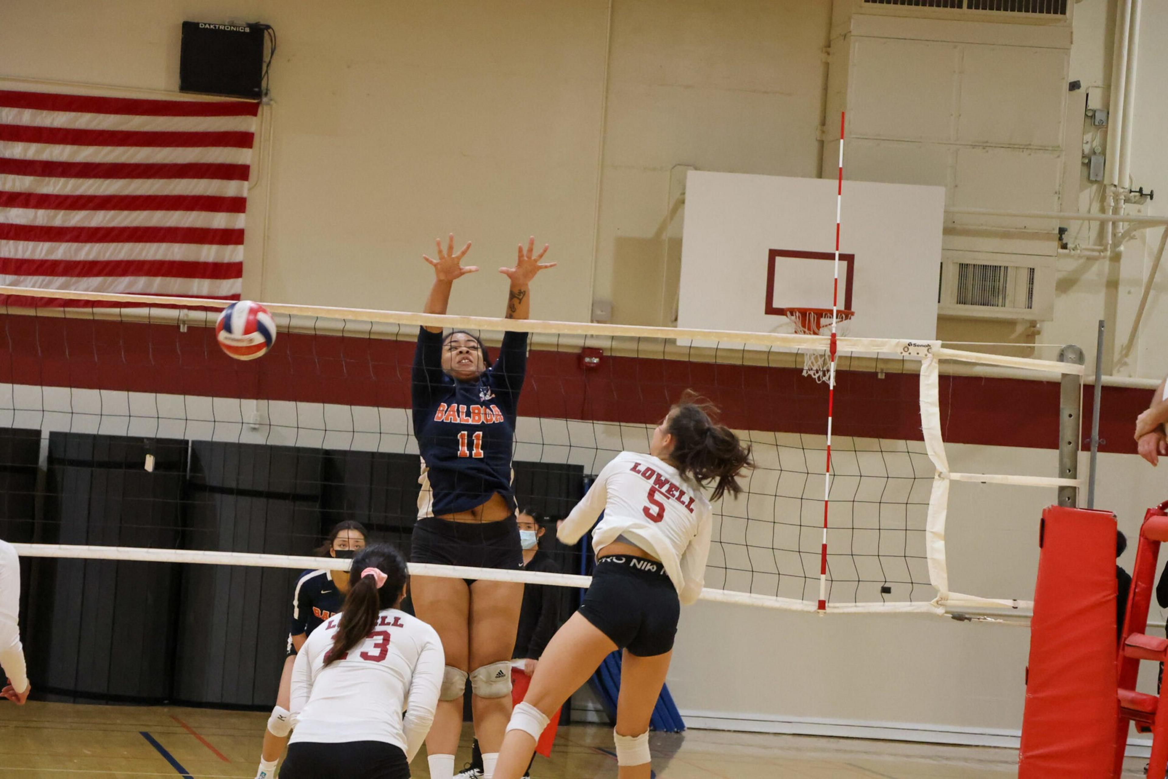 A volleyball player in a black uniform with number 11 jumps to block the ball while a player in a white uniform with number 3 prepares to spike it.