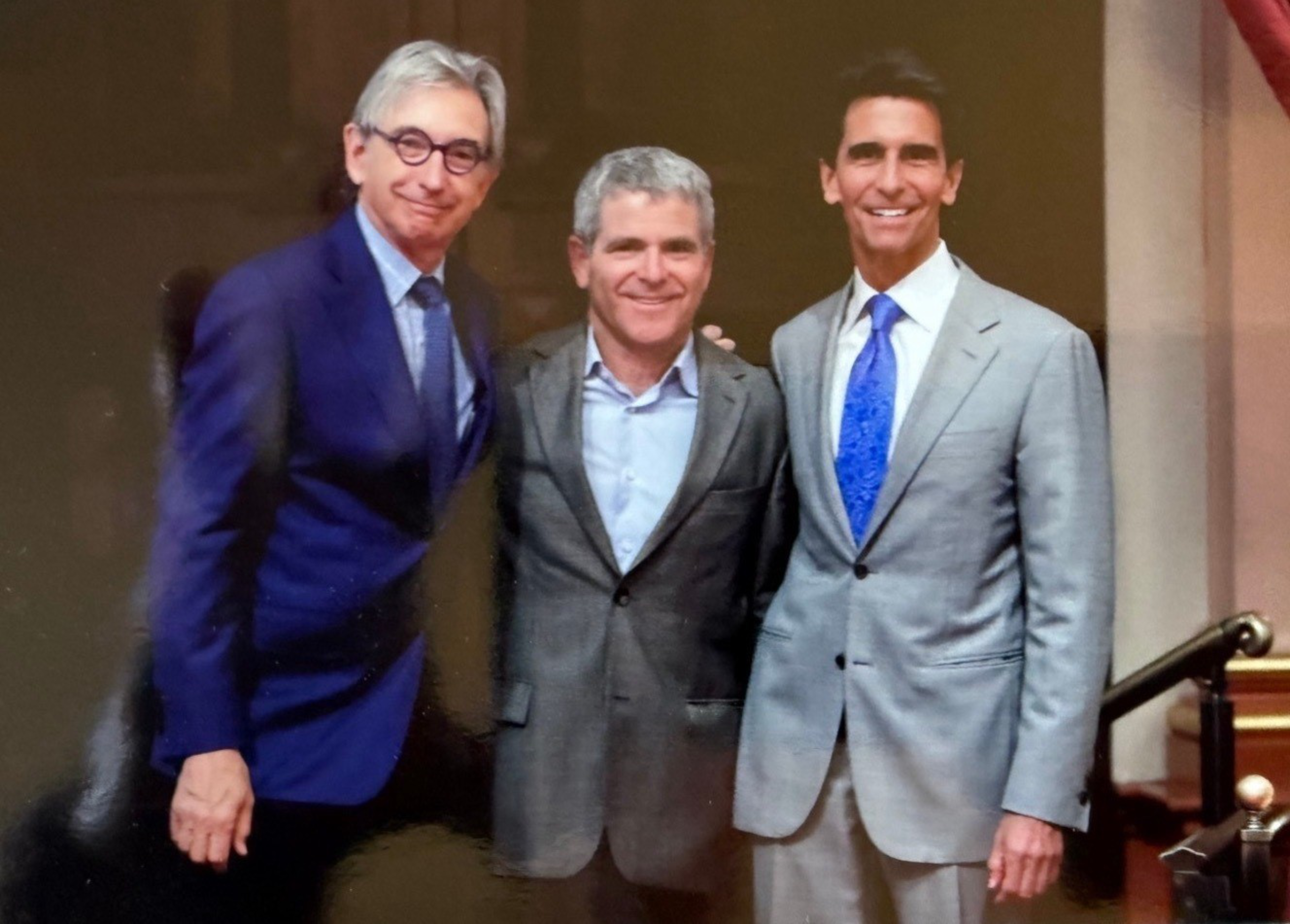 Three men in suits stand close together smiling; the man on the left wears a blue suit and round glasses, the middle one has gray hair and a gray suit, and the right one wears a light gray suit with a blue tie.