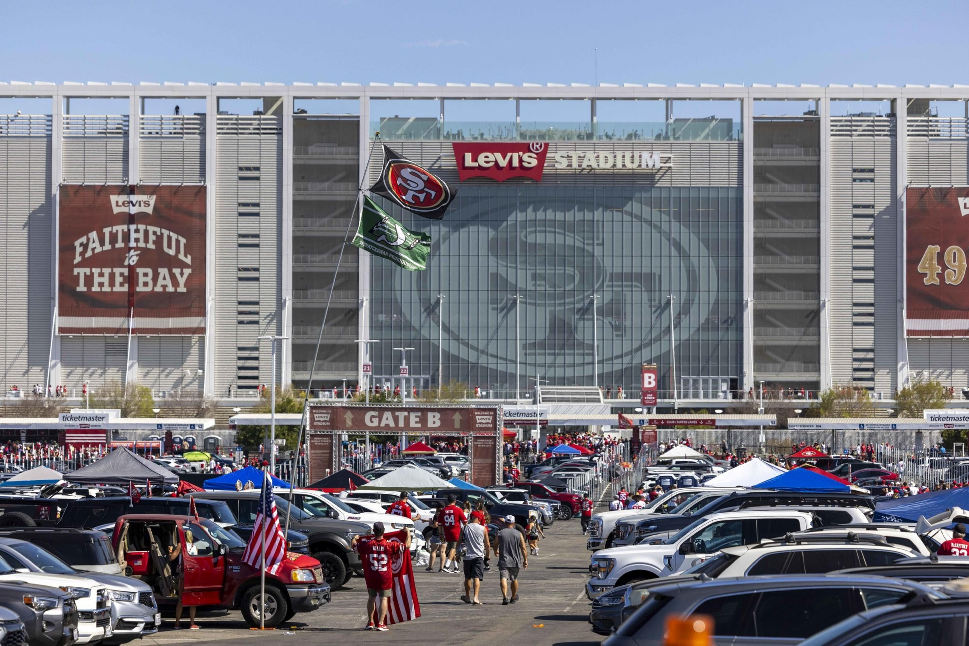 A crowded parking lot outside a stadium features fans tailgating under tents, cars adorned with flags, and a large stadium banner reading &quot;Faithful to the Bay.&quot;
