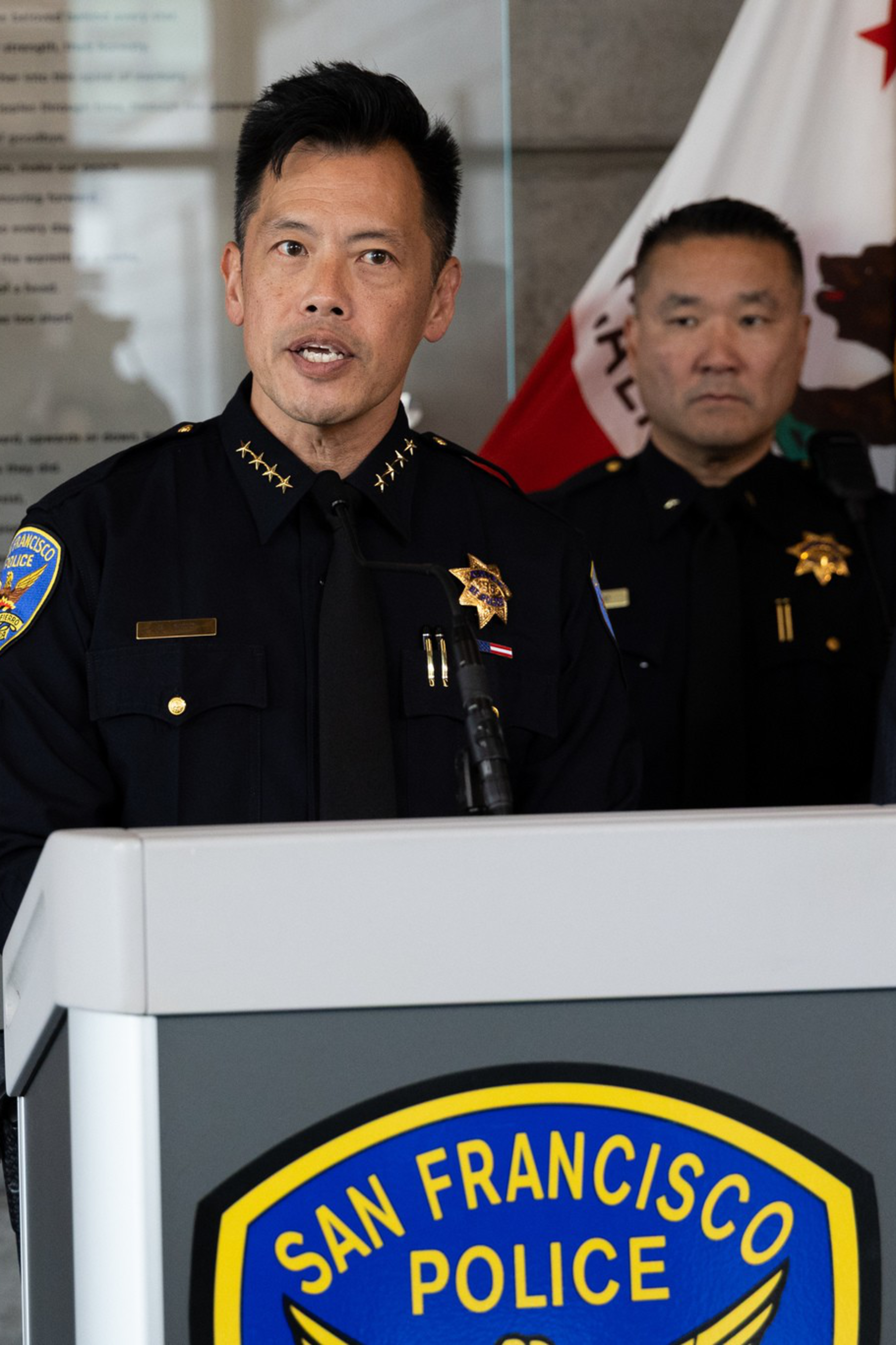 A police officer in uniform speaks at a podium labeled "San Francisco Police" with three formally dressed individuals standing beside him.
