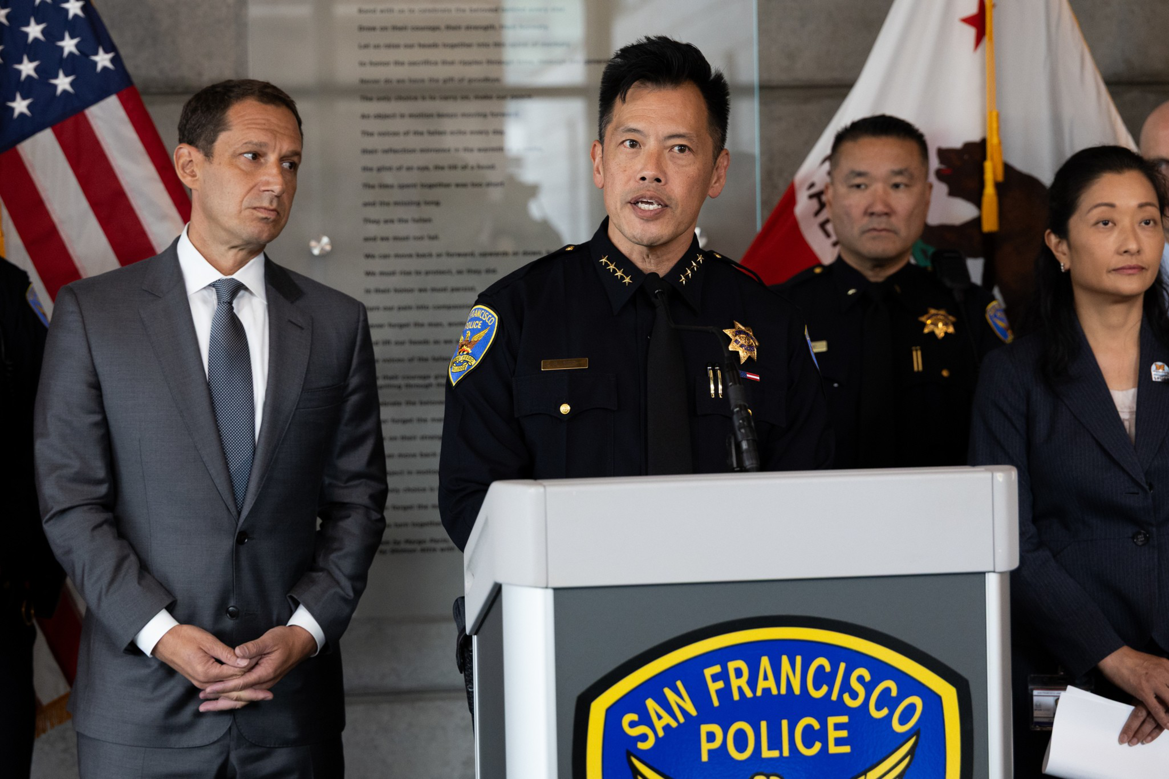 A police officer in uniform speaks at a podium labeled "San Francisco Police" with three formally dressed individuals standing beside him.