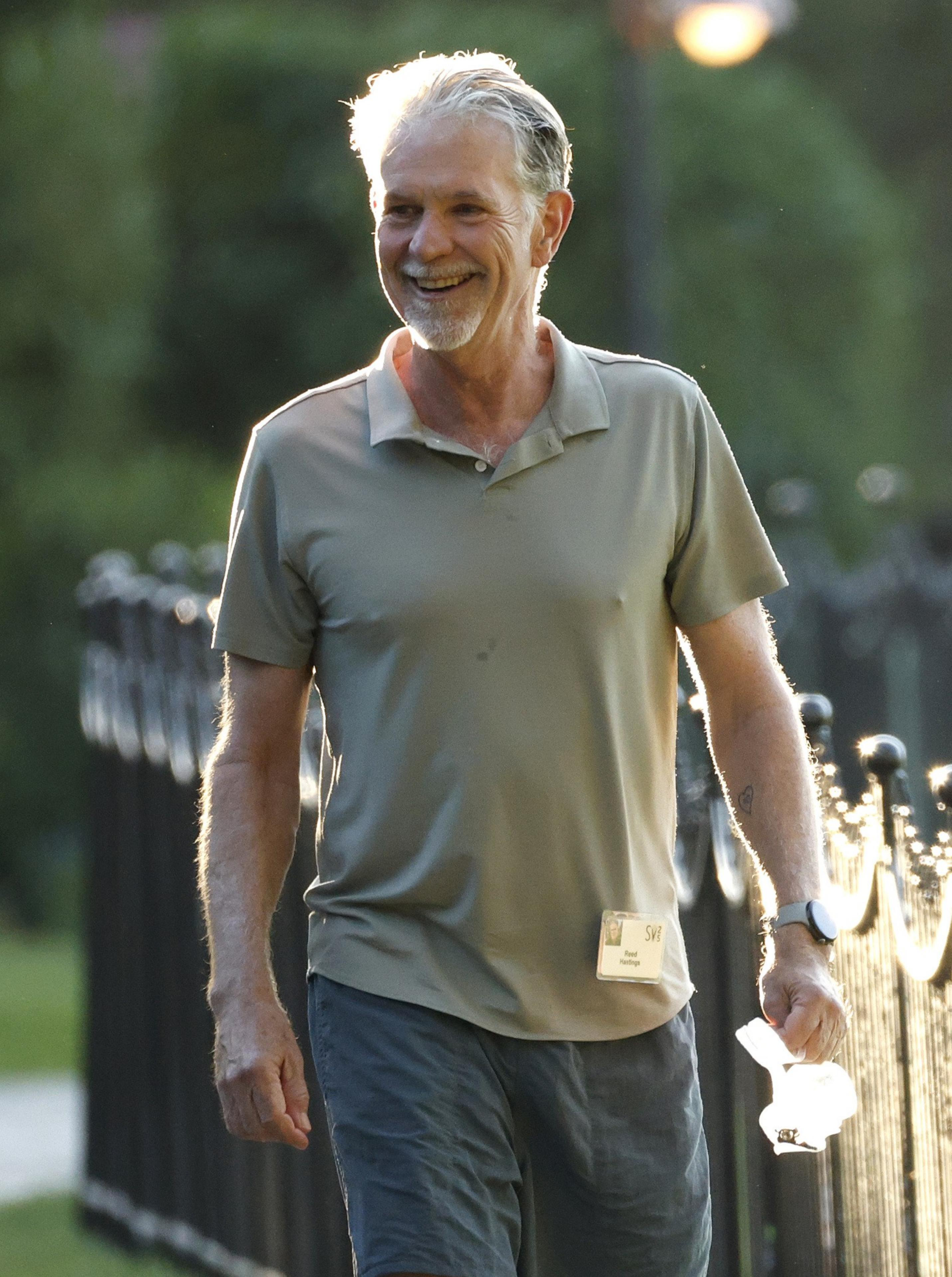 A smiling older man with gray hair and a beard wears a gray polo shirt, dark shorts, and a wristwatch while holding a crumpled paper in one hand.