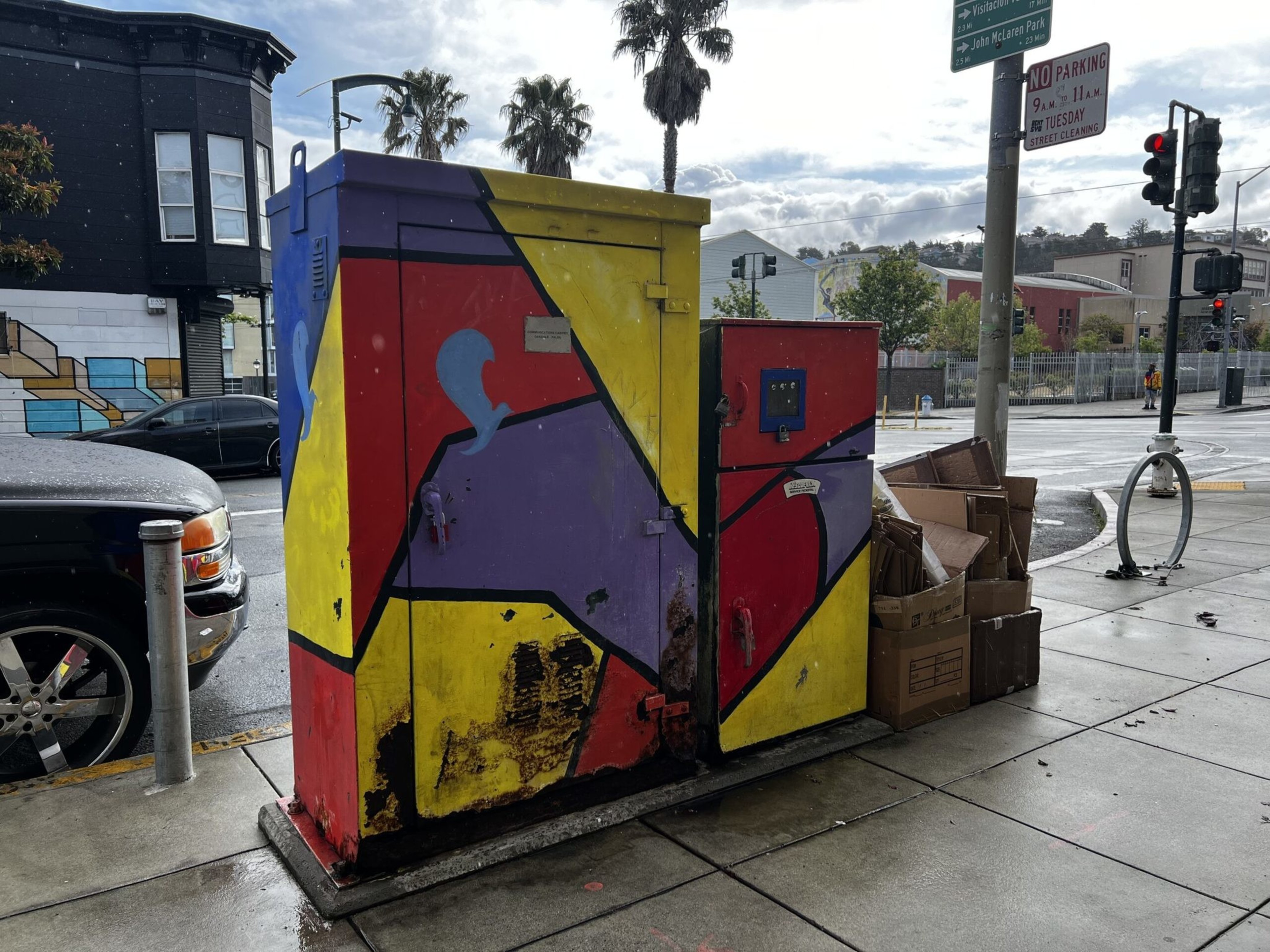A colorful utility box painted with geometric shapes in red, yellow, purple, and blue stands on a wet sidewalk next to stacked cardboard boxes.