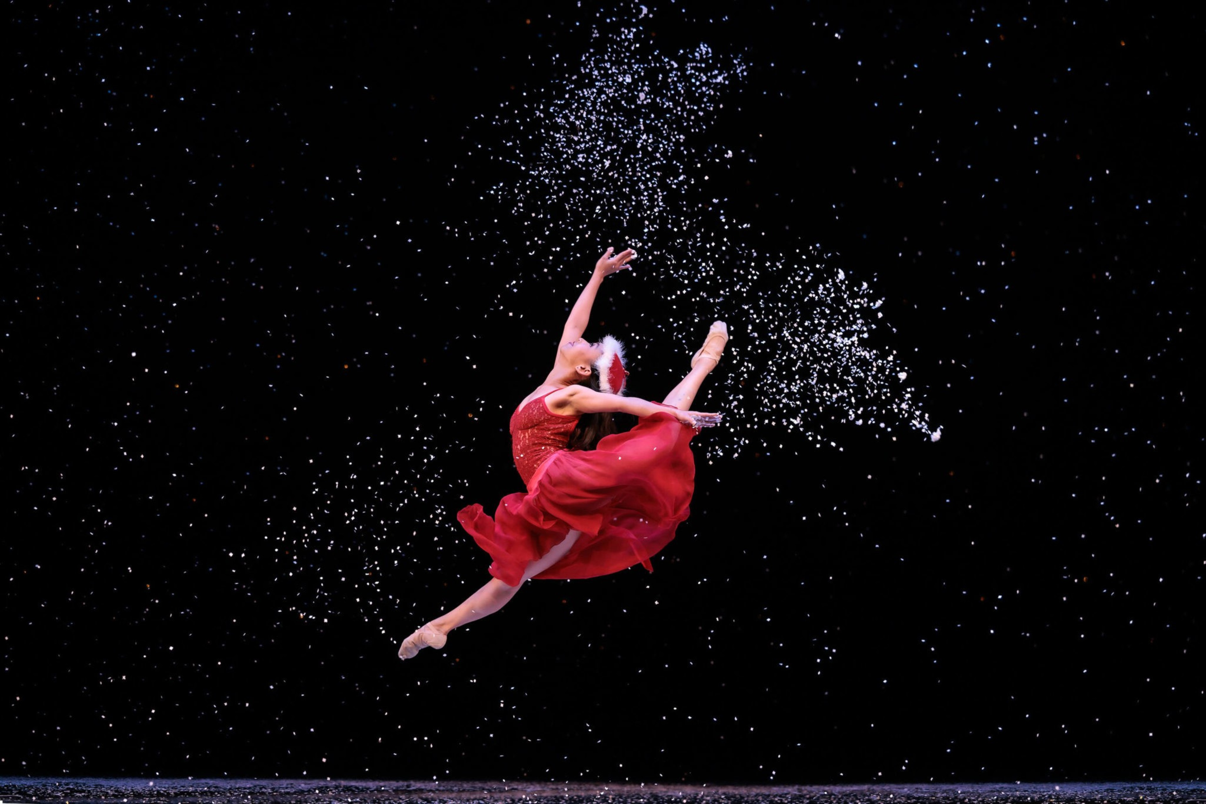 A ballerina in a flowing red dress leaps gracefully mid-air against a black background, with white particles scattered around her like snow.