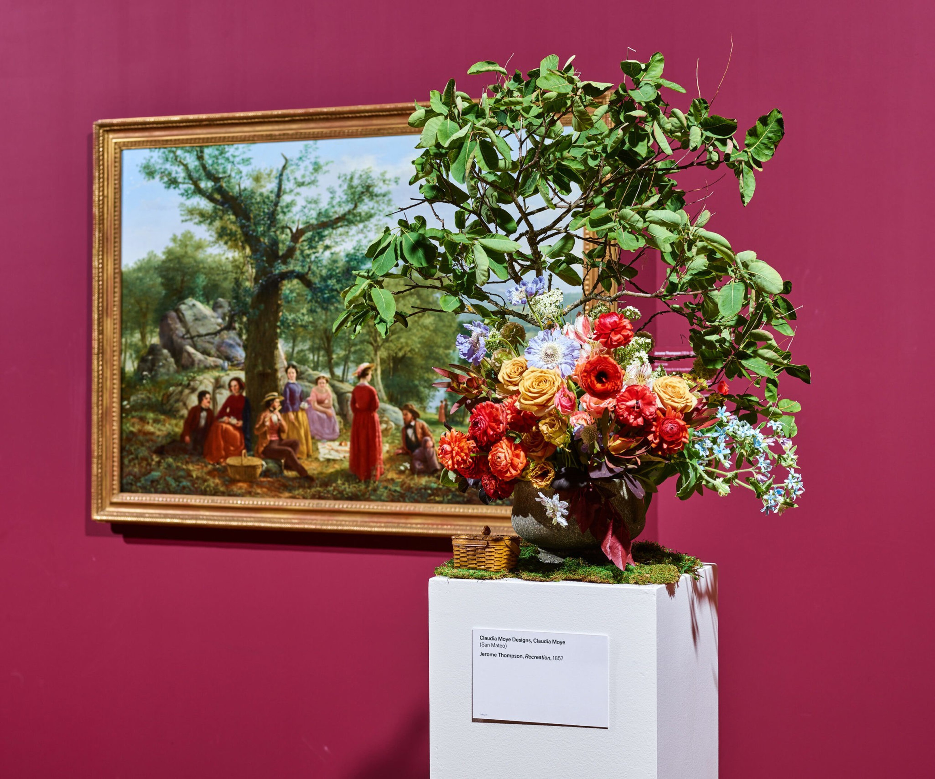 A vibrant flower arrangement with green leaves and red, yellow, and purple flowers sits on a white pedestal in front of a framed painting of people gathered under a tree.