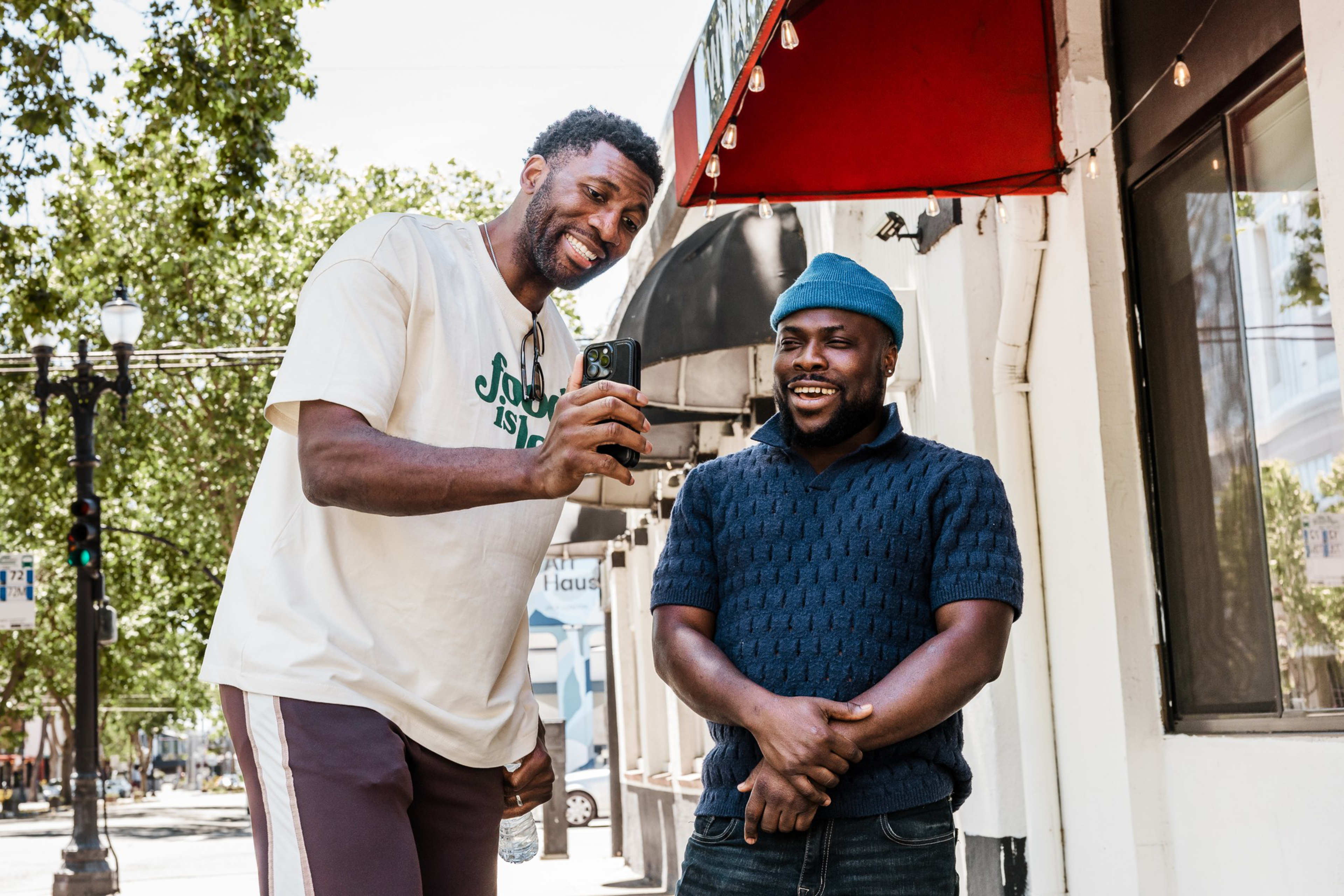 Two men stand outside a building smiling, one wearing a white shirt holding a smartphone showing it to the other in a blue shirt and beanie.
