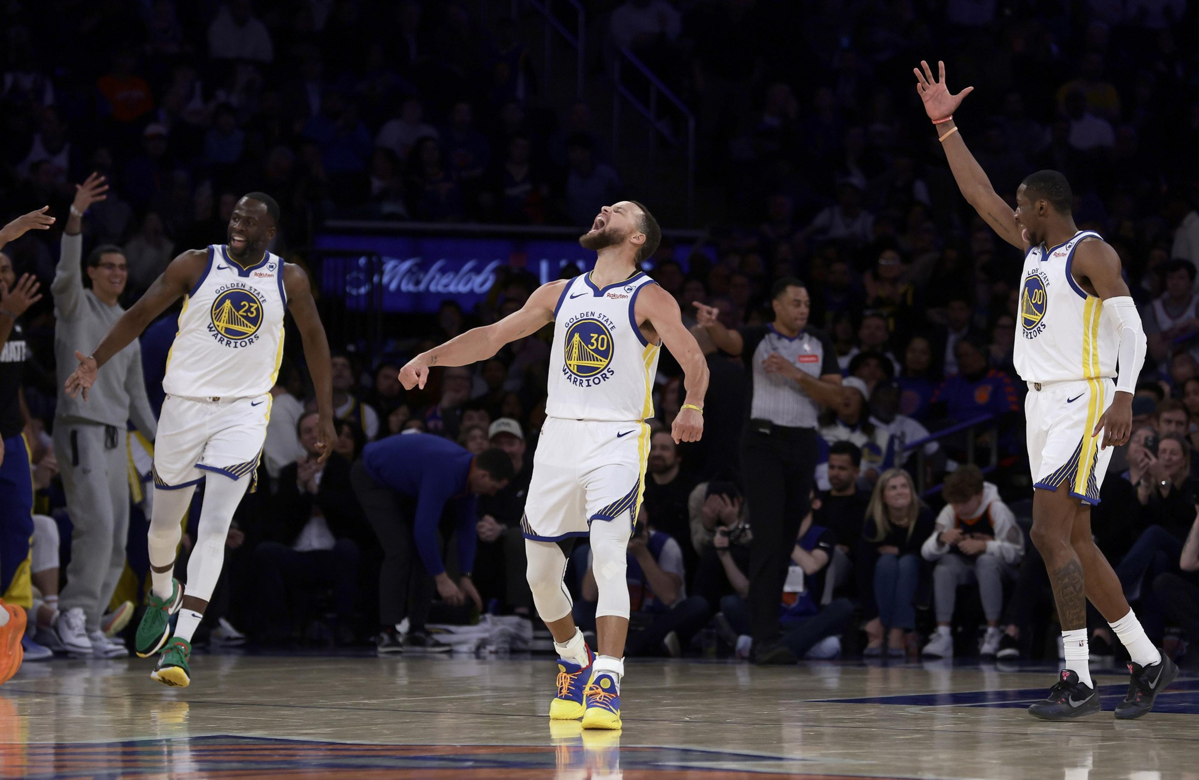 Three basketball players, wearing Golden State Warriors jerseys, celebrate energetically on the court as an official stands nearby and a crowd watches in the background.