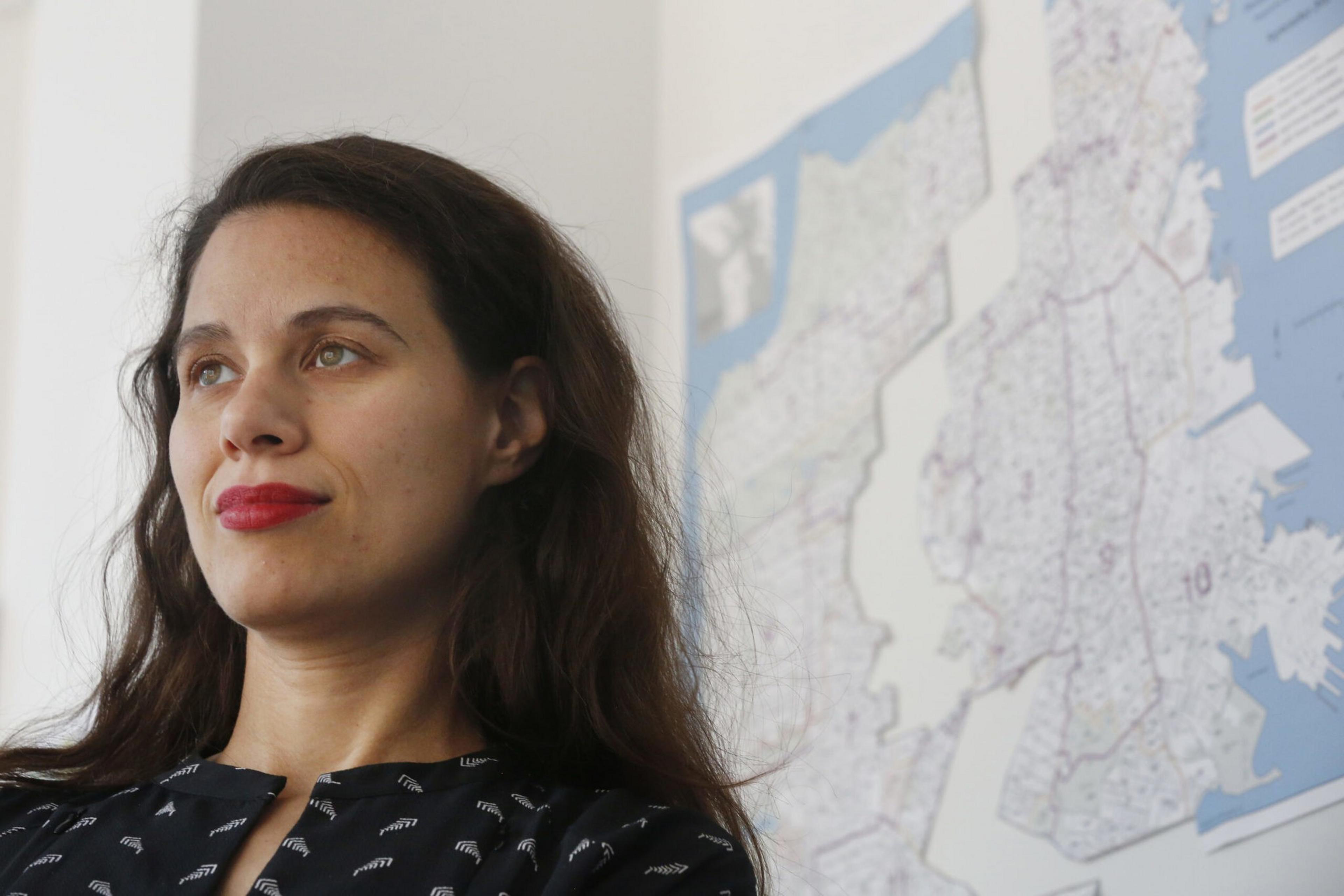 A woman with long dark hair and red lipstick looks slightly upward, standing near a wall with a large, detailed map behind her.