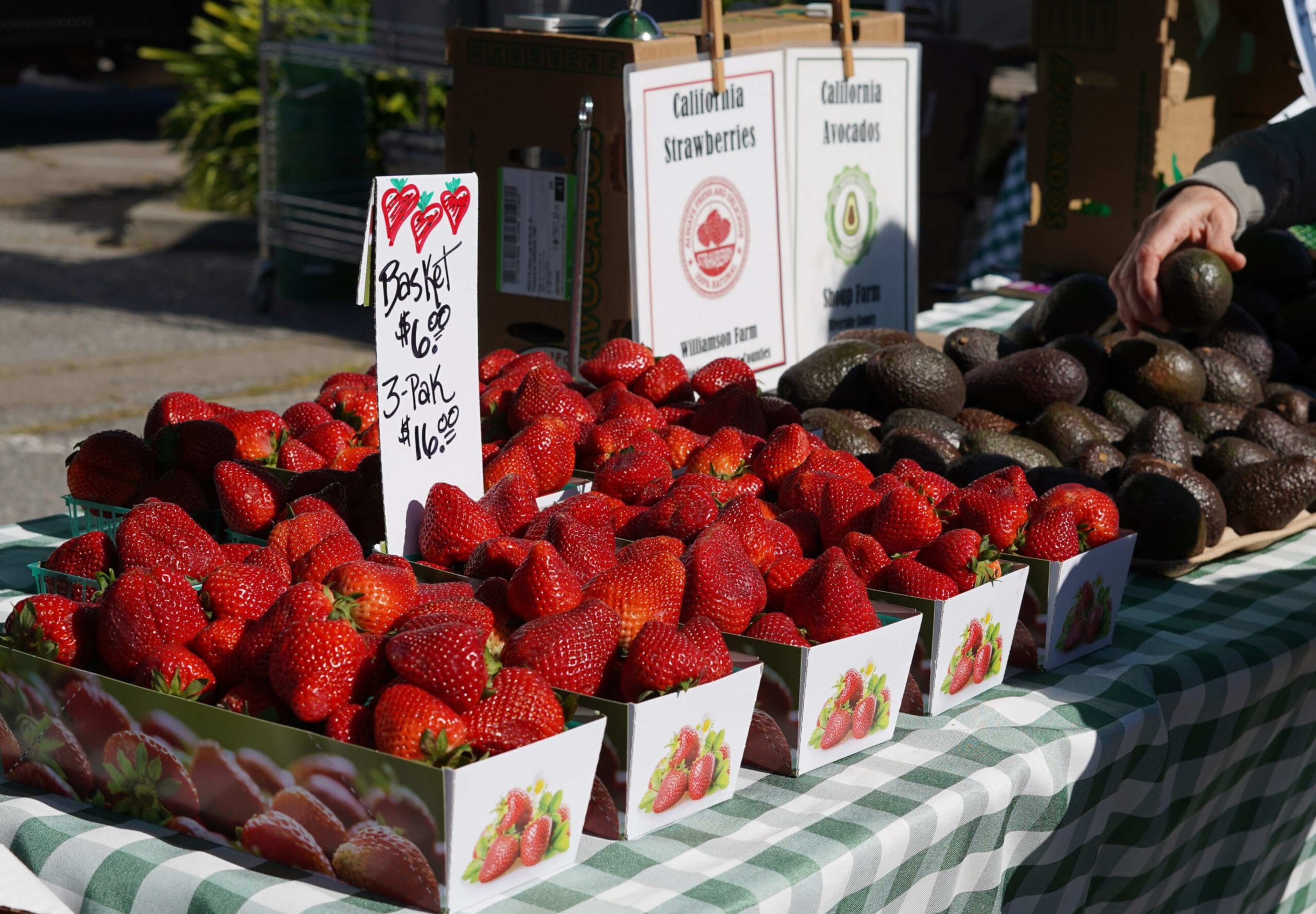A farmers market stand displays baskets of bright red strawberries with a price sign. Nearby, there are tables with avocados. Green checkered cloth covers the tables.