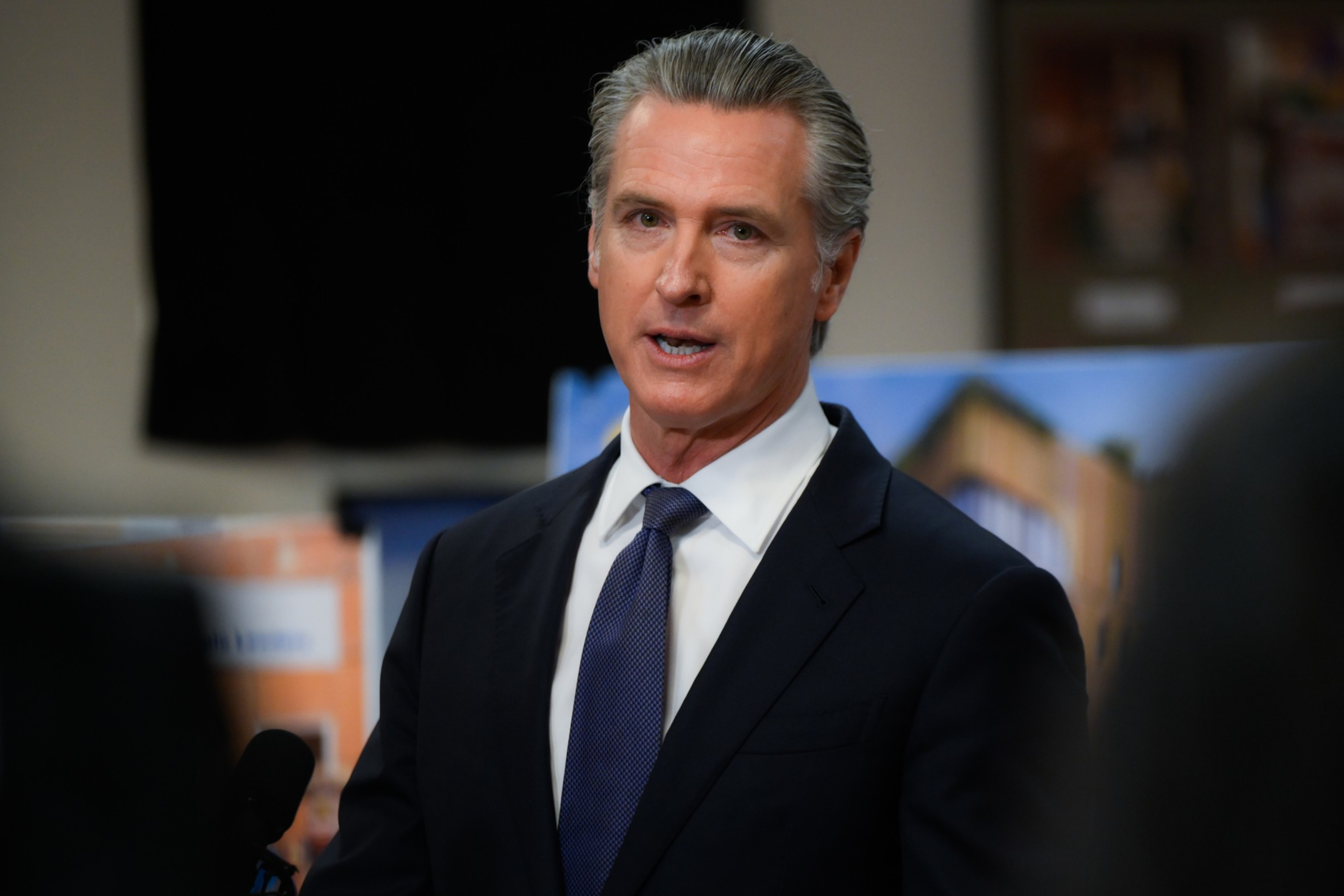 A man with gray hair, wearing a dark suit, white shirt, and blue tie, speaks in front of blurred background posters and a microphone.