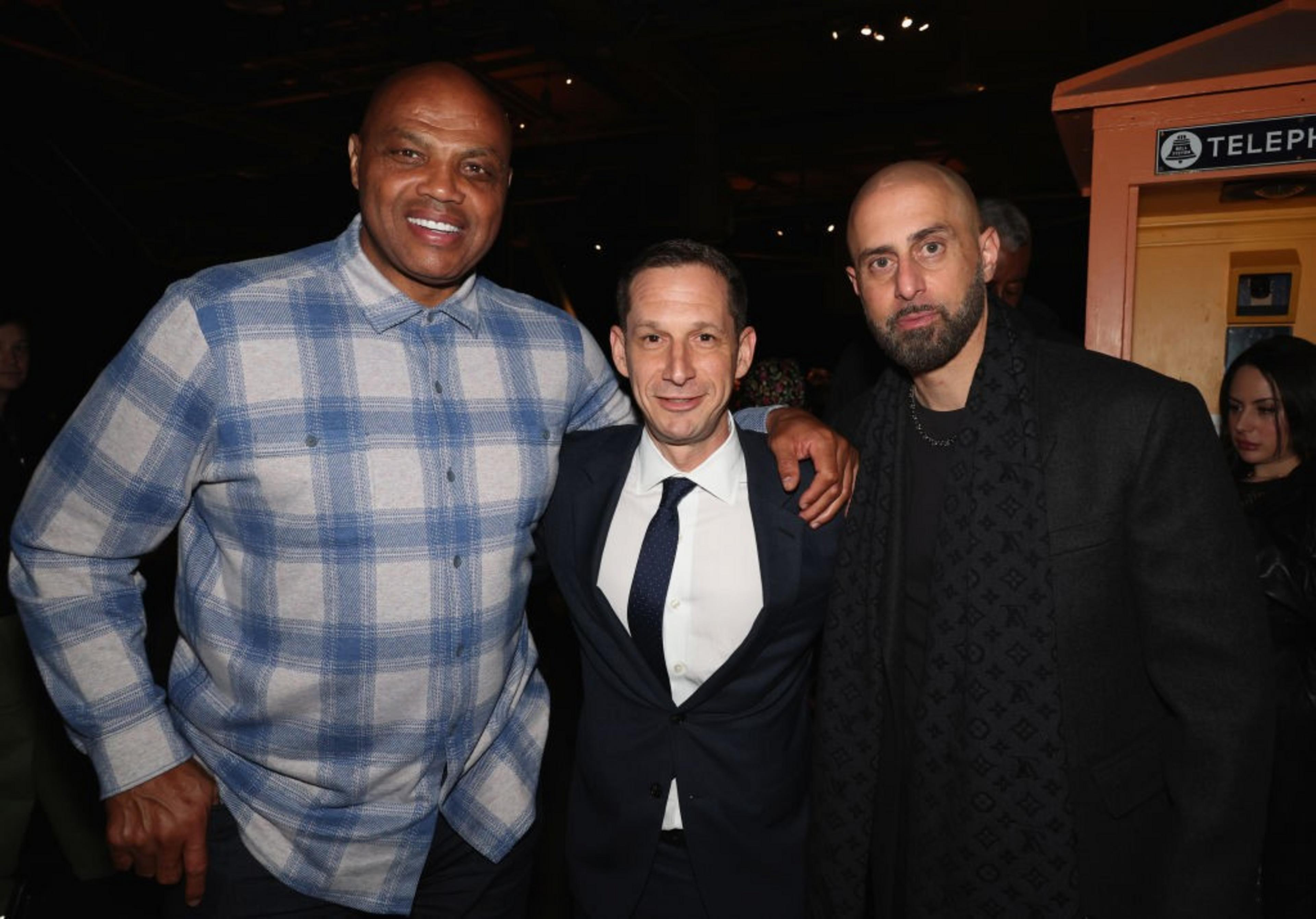Three men are standing close together, smiling. The man on the left wears a blue plaid shirt, the middle one is in a suit, and the man on the right is in a dark coat.