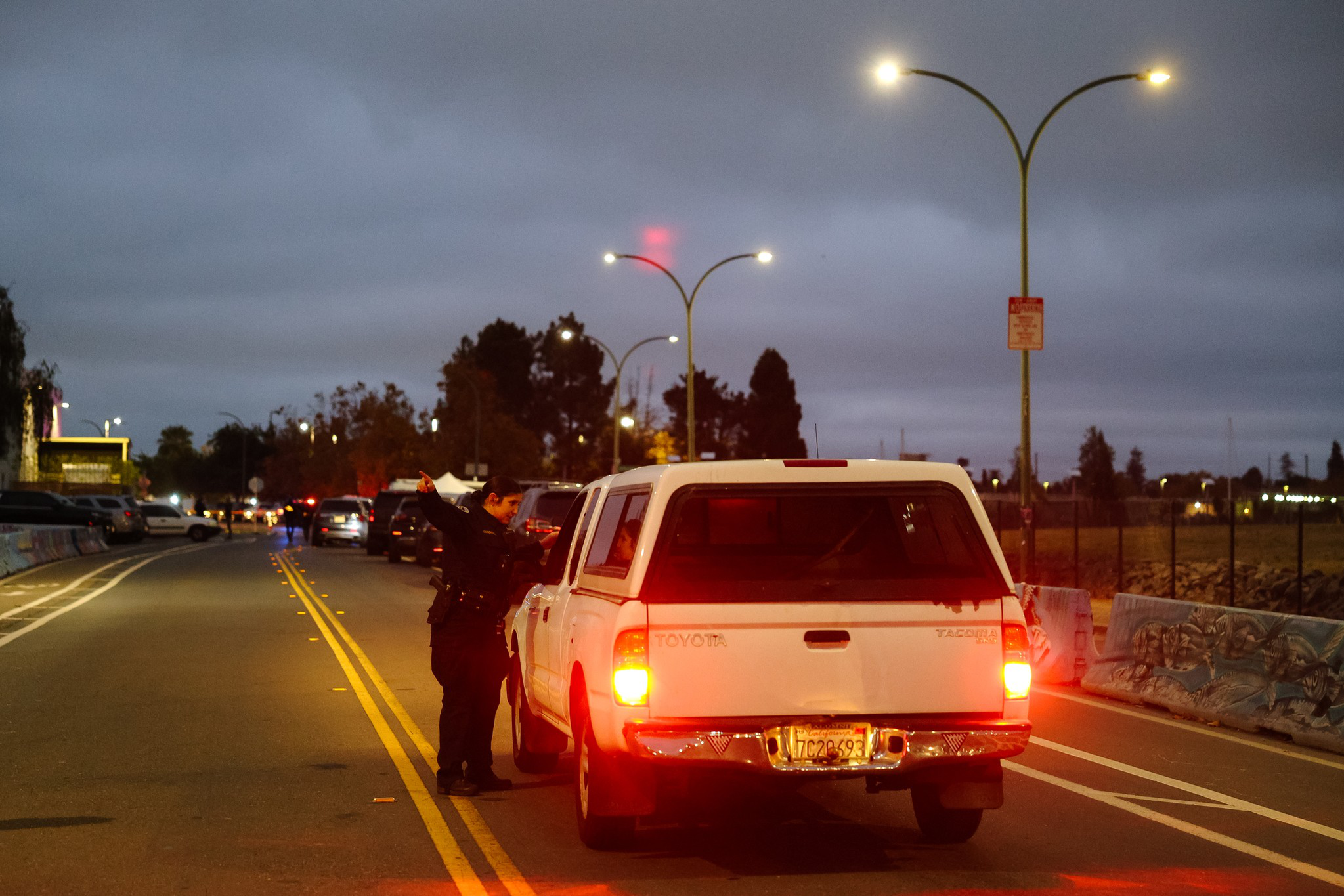 A police officer is speaking to the driver of a white Toyota Tacoma during a traffic stop on a dimly lit road at dusk.