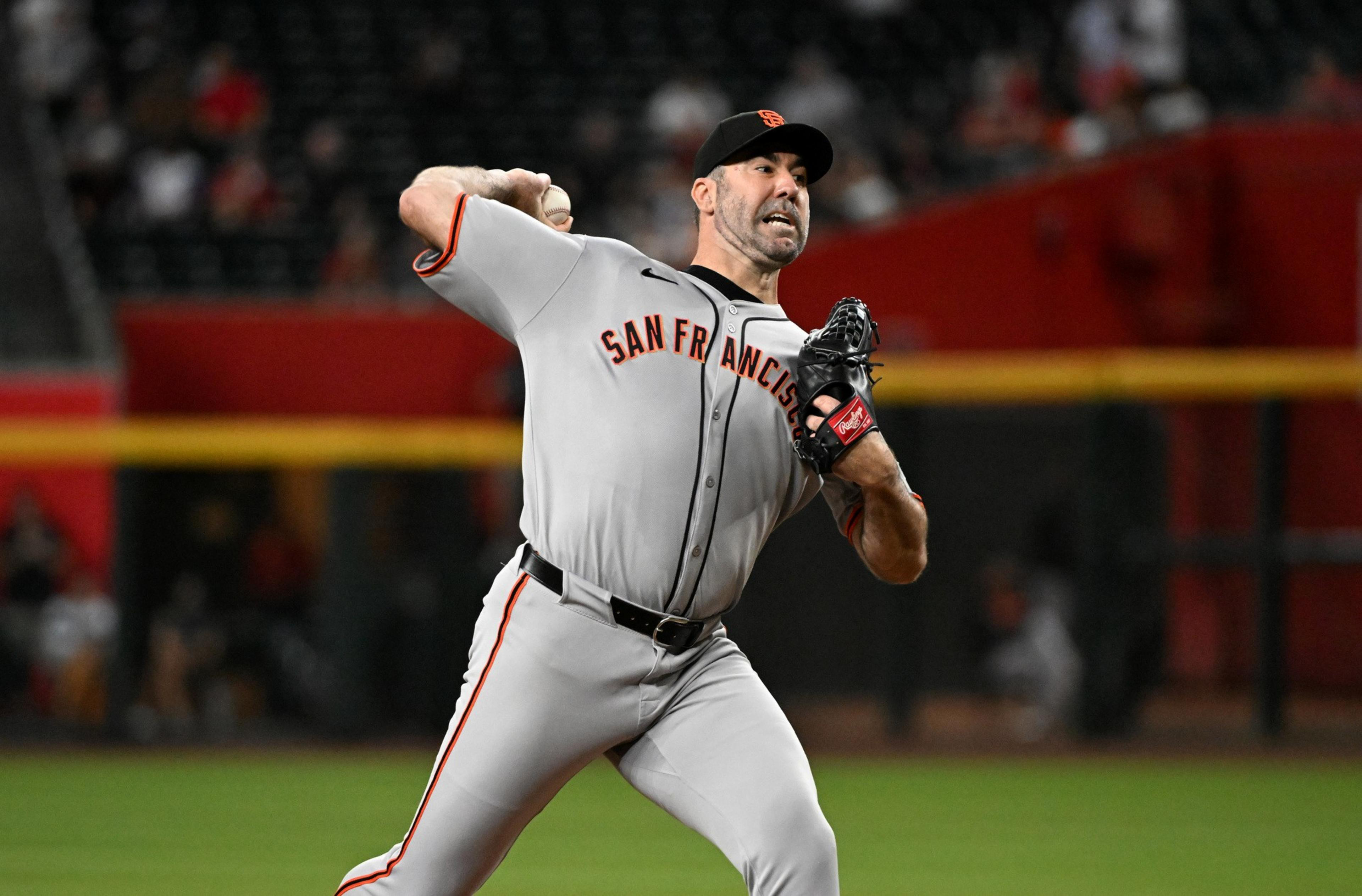 A San Francisco Giants pitcher is in mid-throw, wearing a gray uniform and black cap, on a baseball field with blurred spectators in the background.