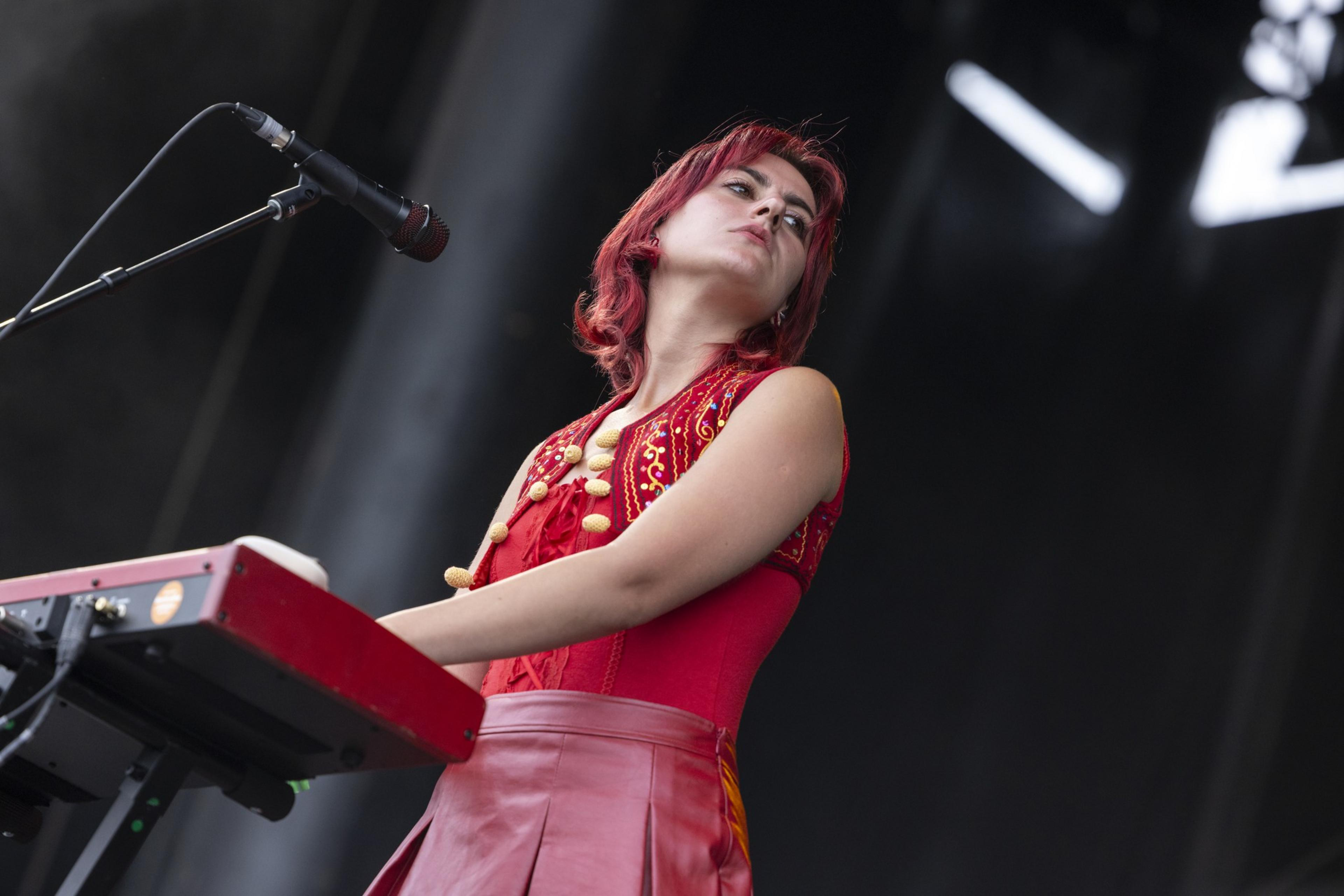A person with red hair, wearing a red outfit, stands at a keyboard on stage, looking up with a serious expression. A microphone is positioned near their face.