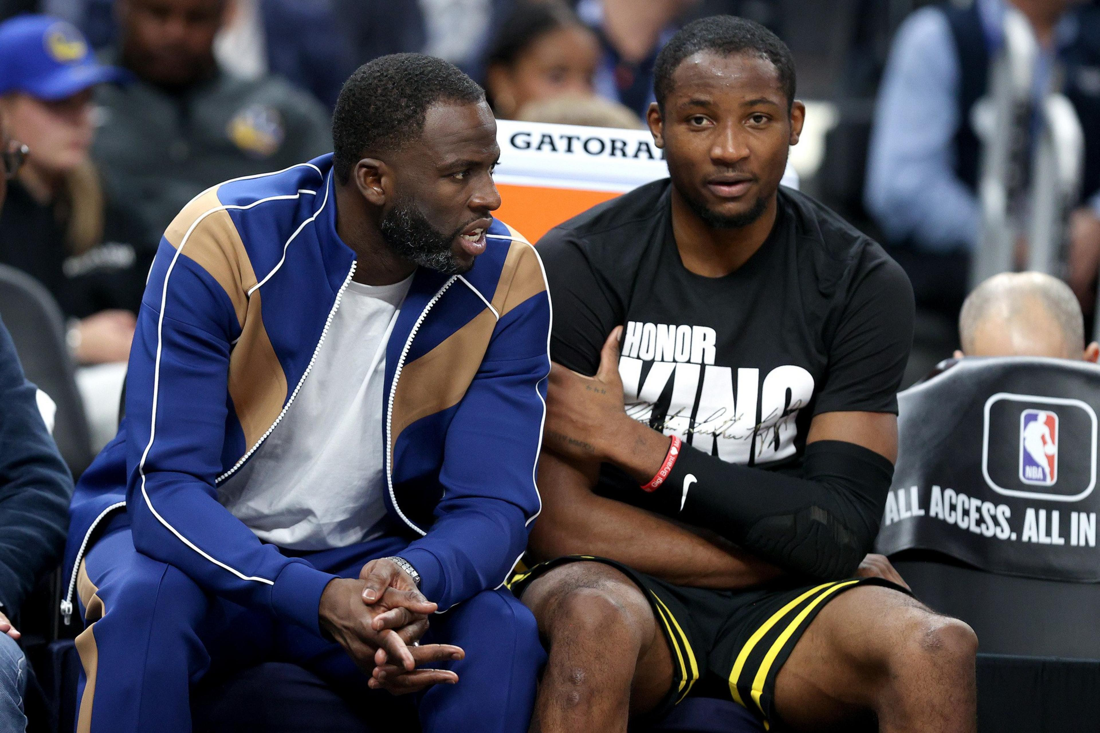 Two men sit on a sports bench. One wears a blue and tan tracksuit, talking animatedly. The other in a black &quot;Honor King&quot; shirt listens quietly.