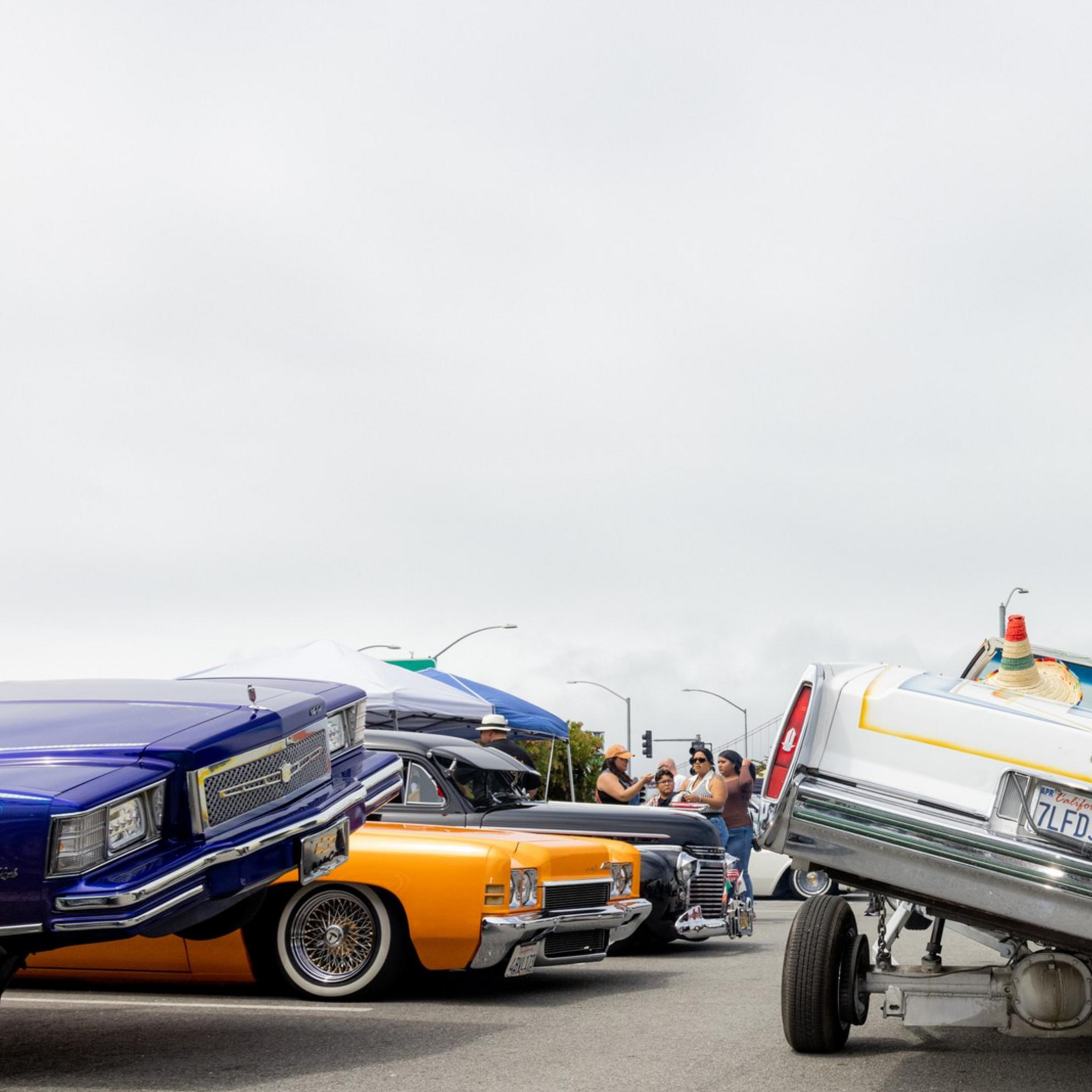 The image shows a white car performing a dramatic hydraulic lift stunt, tilted on one side. Nearby, colorful lowrider cars are parked, and people are watching and taking photos.
