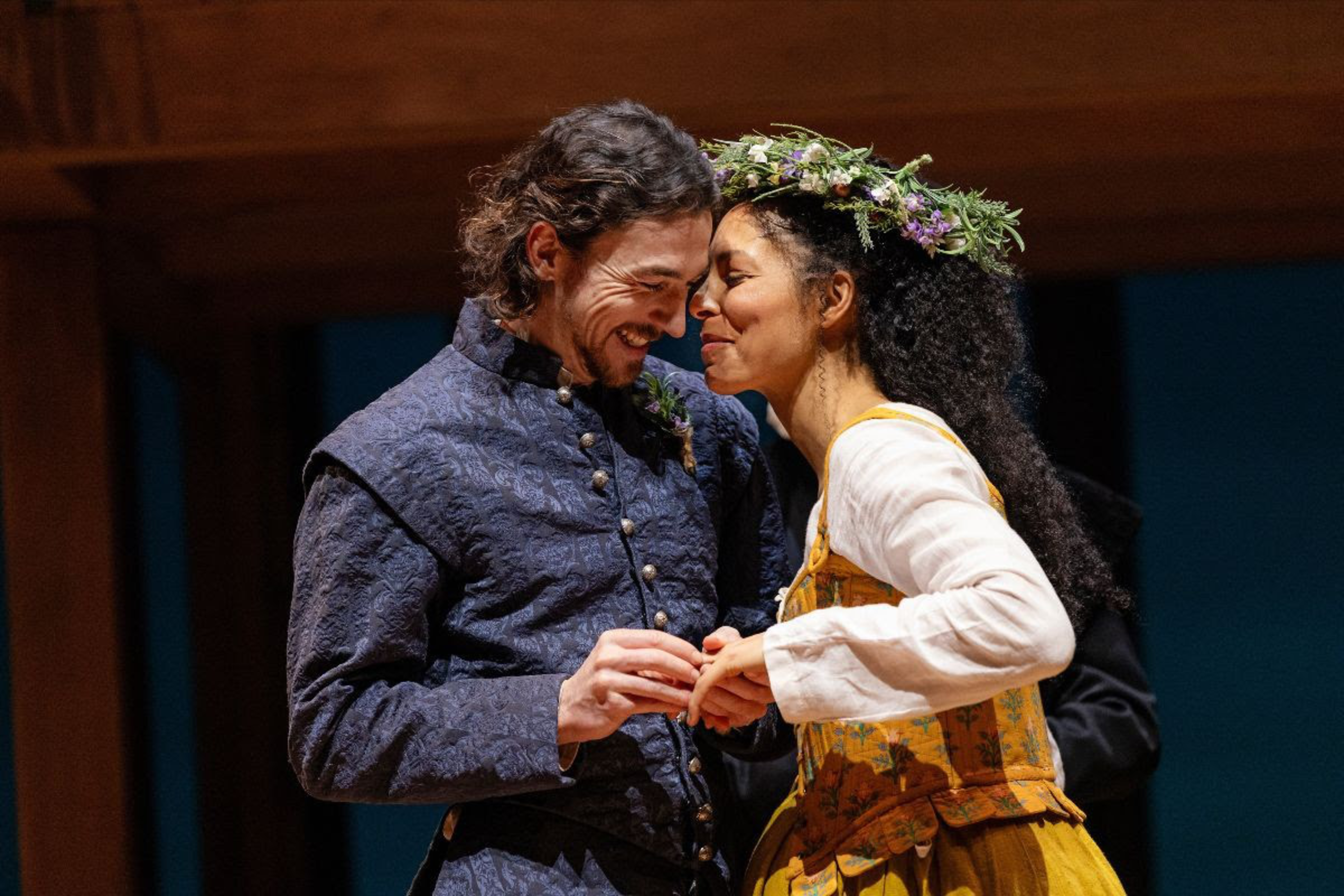 A couple dressed in historical costumes hold hands closely, smiling warmly, with the woman wearing a flower crown and a yellow dress.