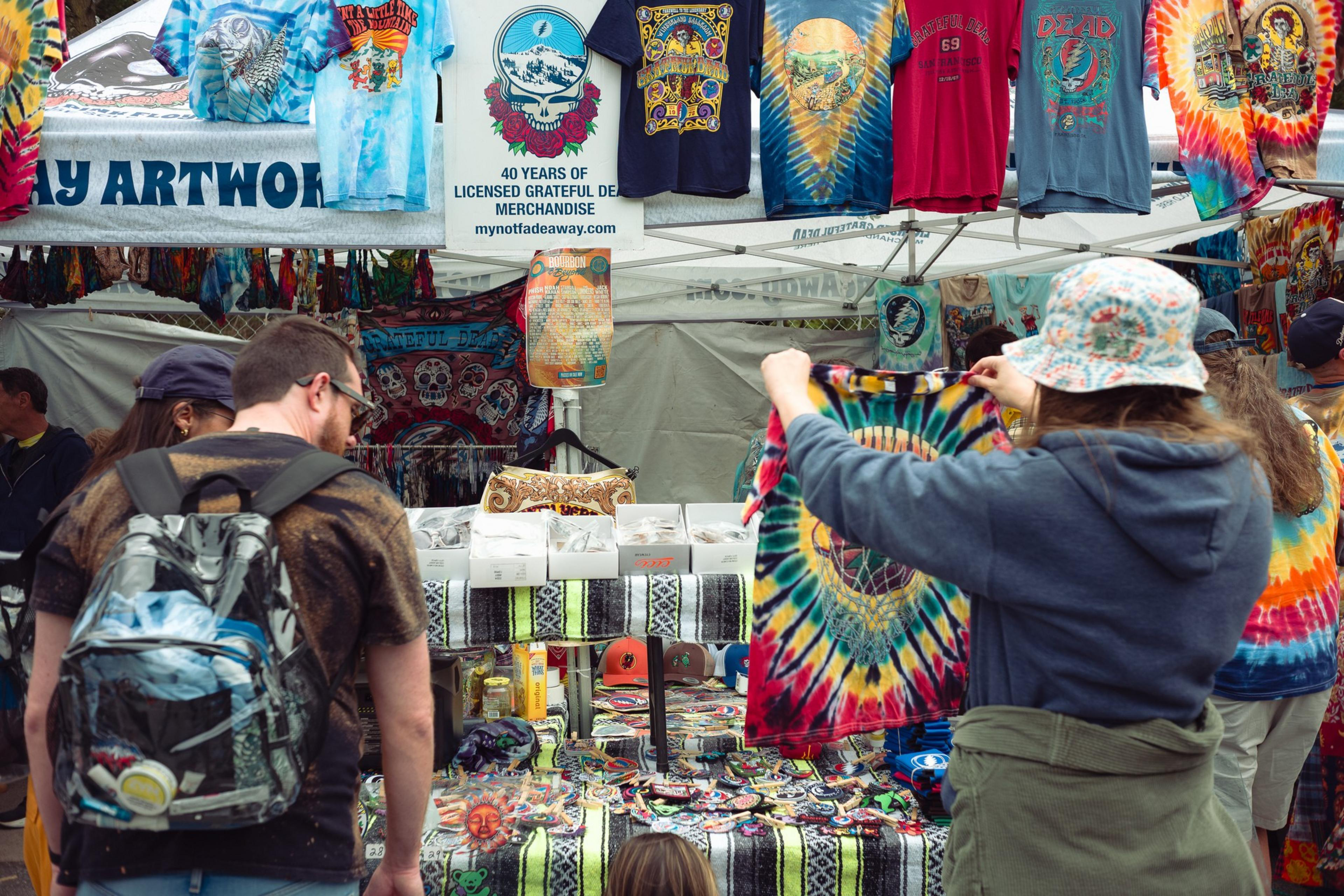People browse a market stall filled with colorful tie-dye and Grateful Dead merchandise, including T-shirts, patches, and accessories.