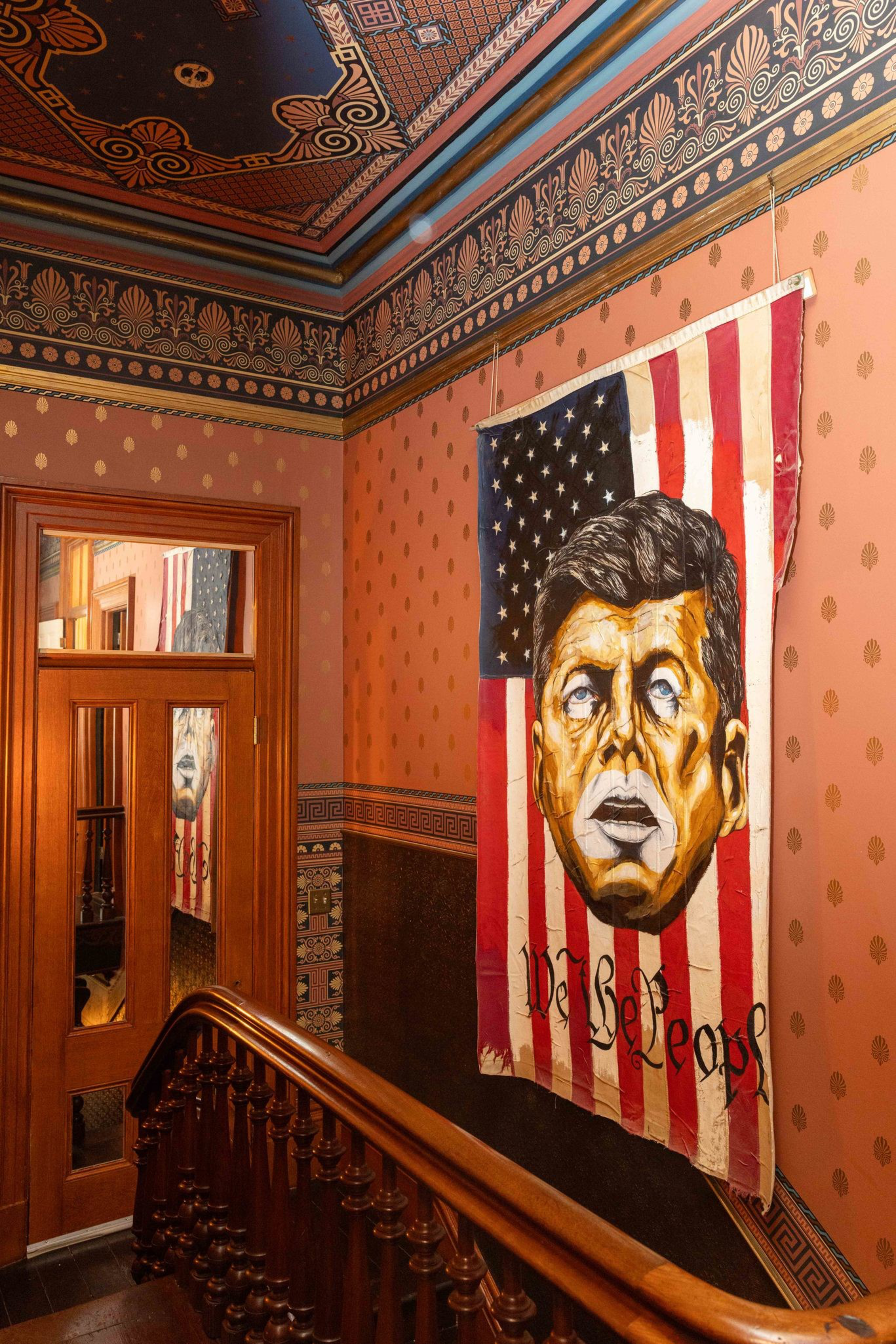 A painted American flag with John F. Kennedy’s face and “We the People” written at the bottom hangs on an ornately decorated wall by a wooden staircase.