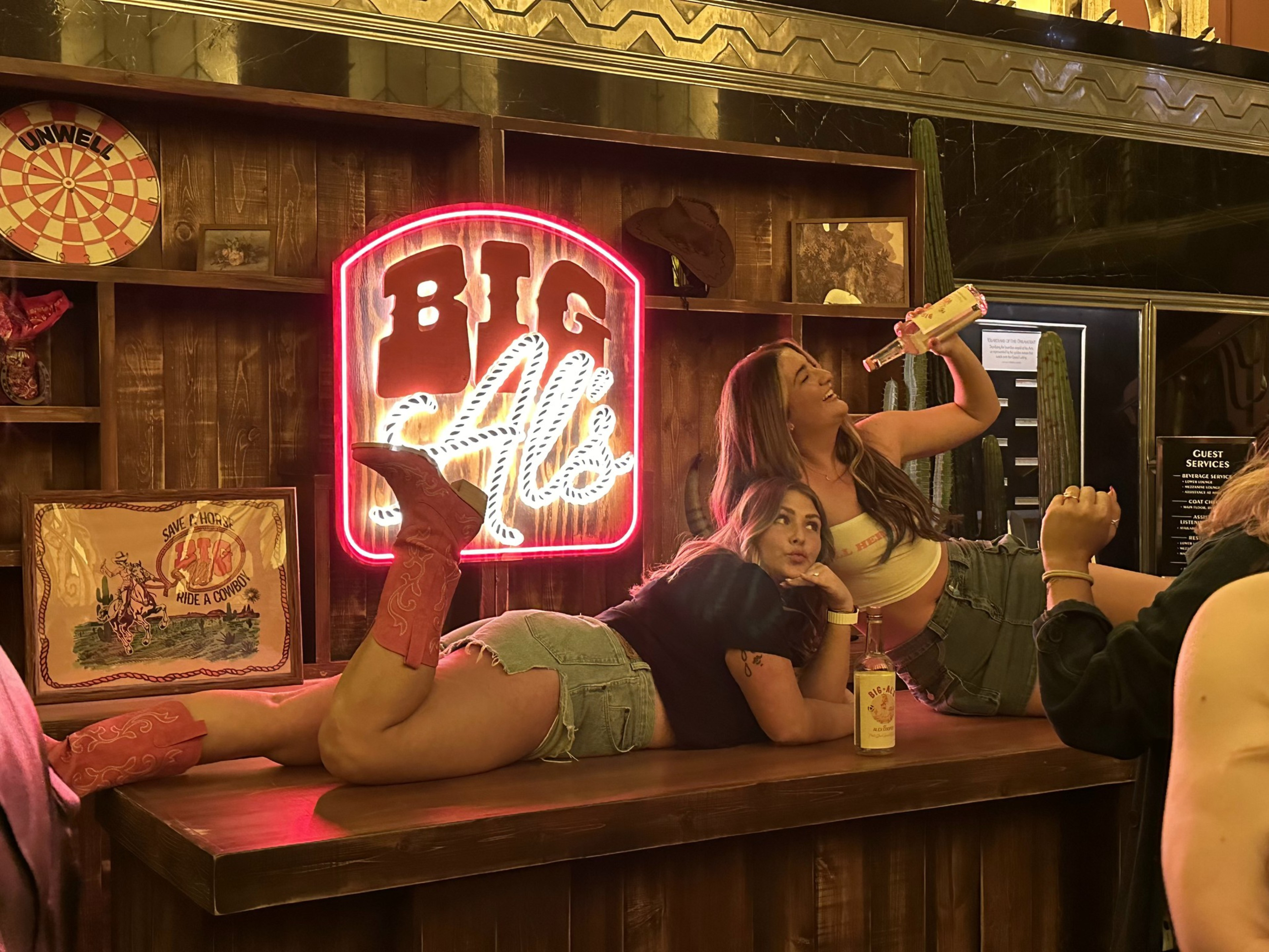 A woman in jean shorts and red cowboy boots and a friend in a jean skirt and a white shirt pose on top of a bar in front of a sign reading &quot;Big Al's.&quot;